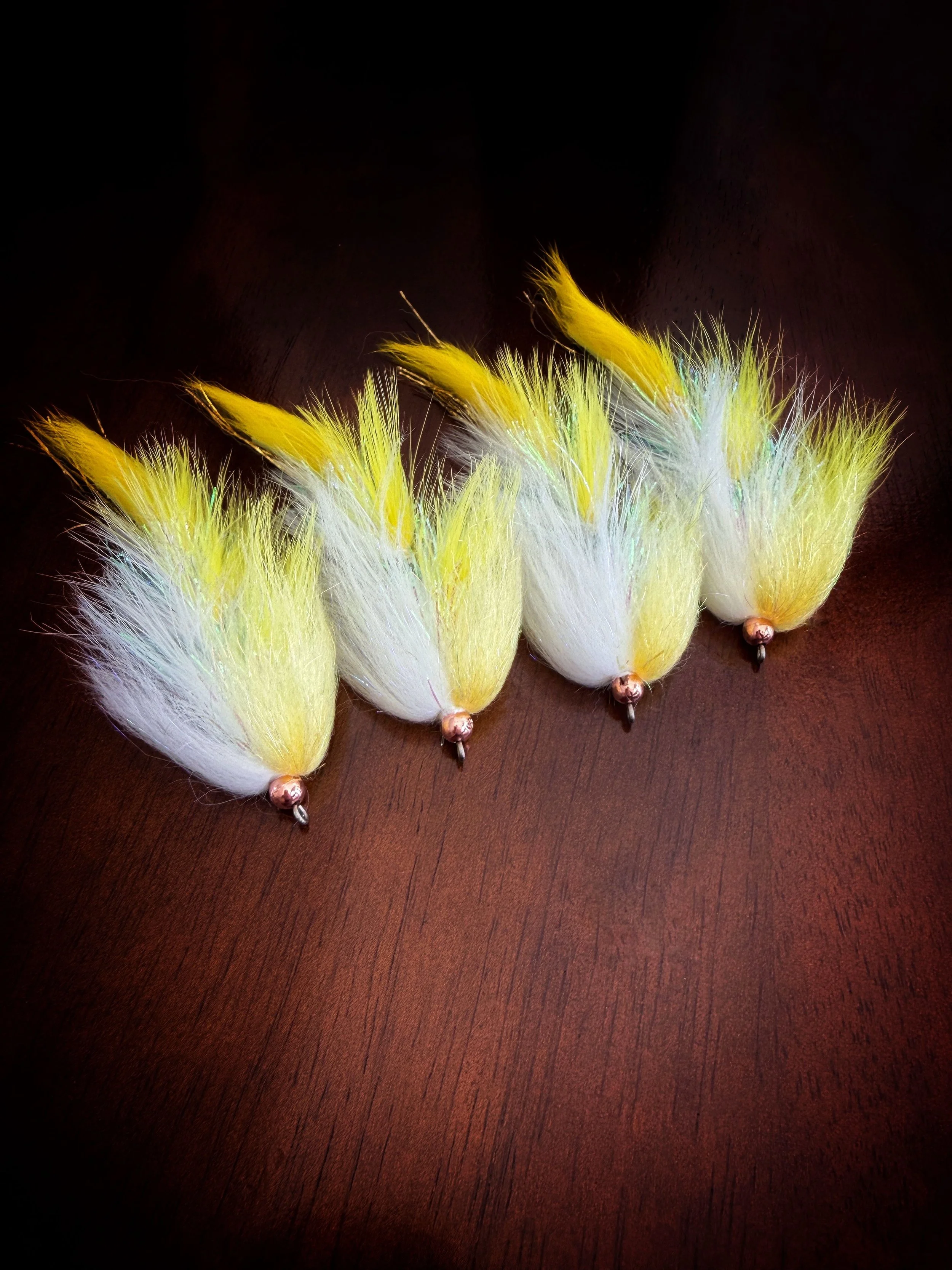 Five yellow and white fluffy fishing lures with metallic hooks, arranged in a row on a dark wooden surface.