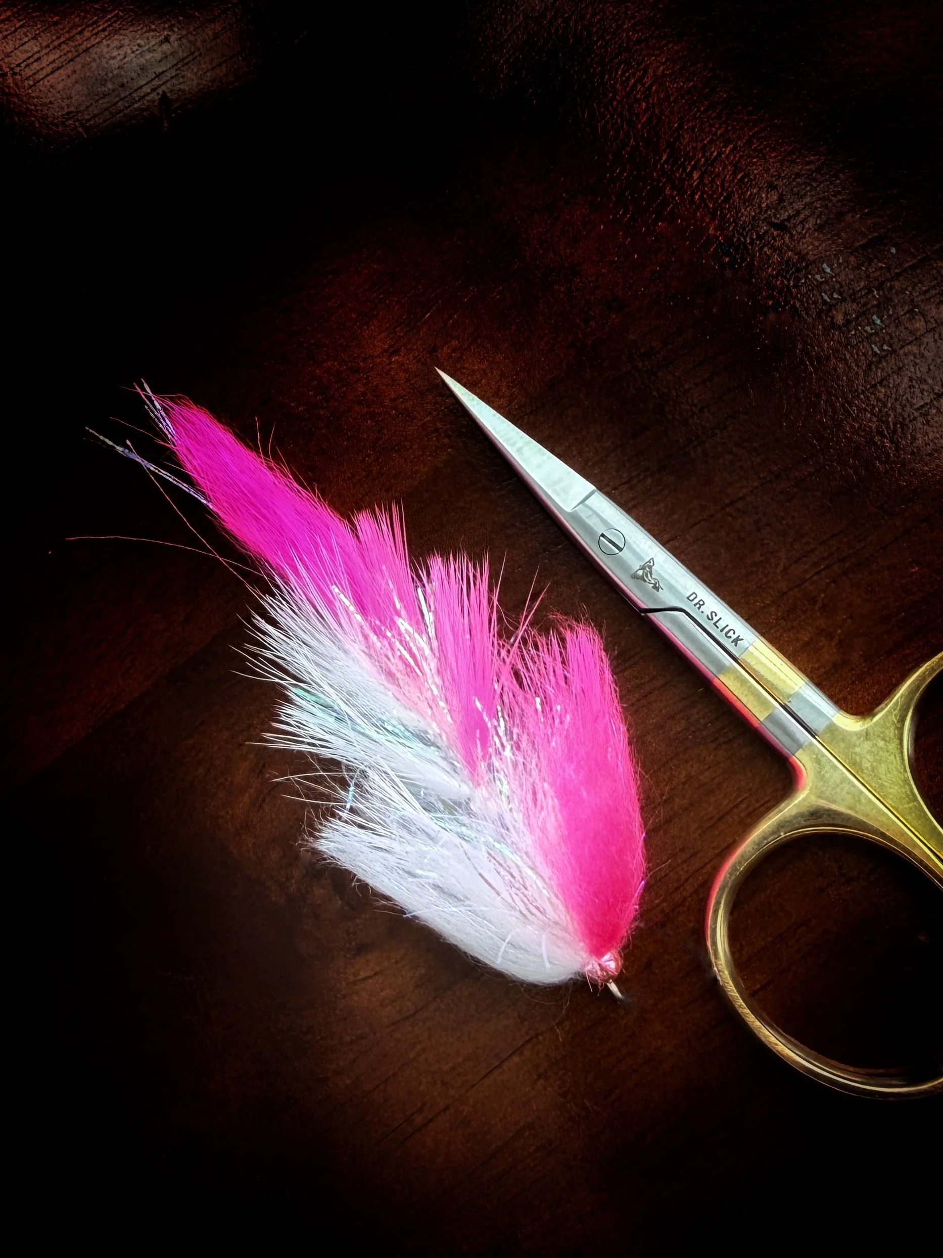 A pink and white feather tied to a metal fishing lure with a sharp blade and gold handle on a dark wooden surface.