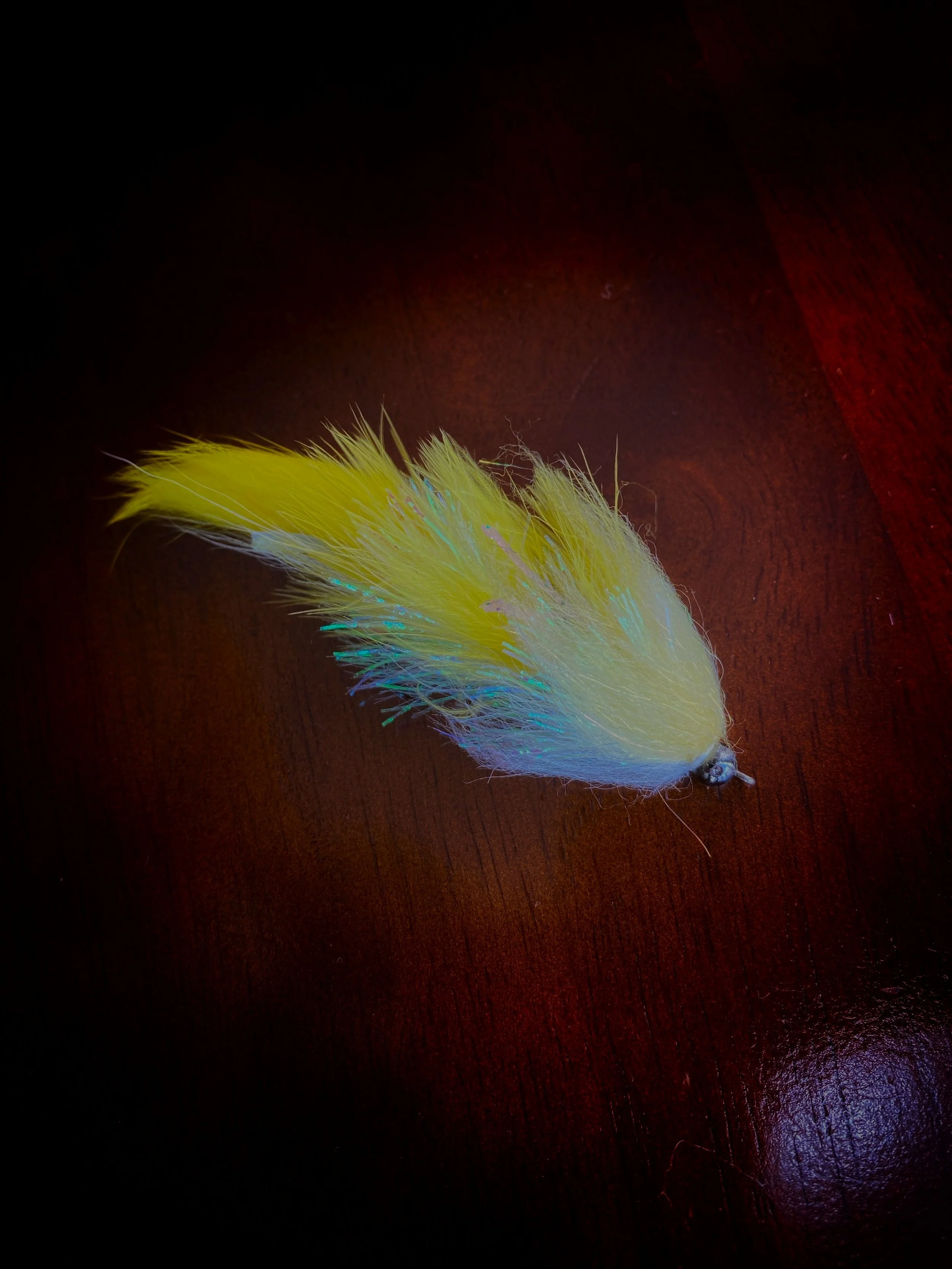 A colorful fishing lure with yellow, white, and iridescent feathers on a dark wooden surface.