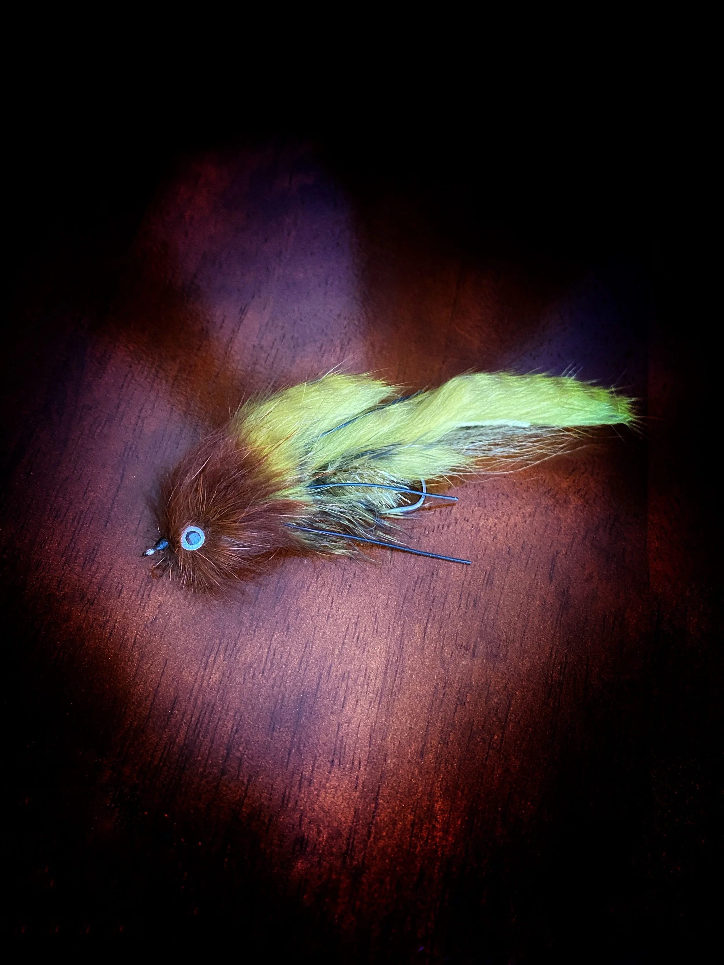 Fishing lure with brown, yellow, and green feathers on a dark wooden surface.
