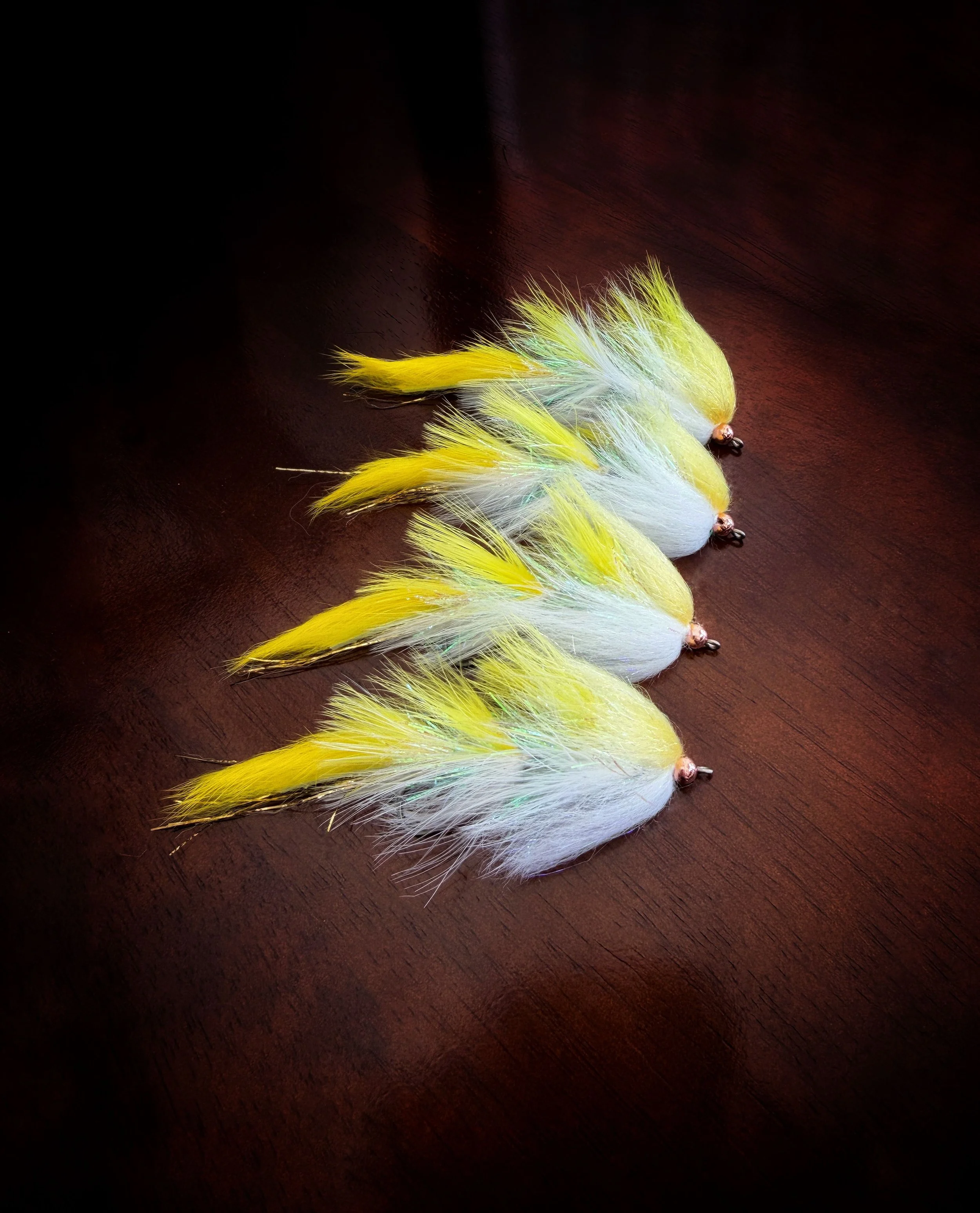 Five fishing lures with yellow and white feathers arranged in a row on a dark wooden surface.
