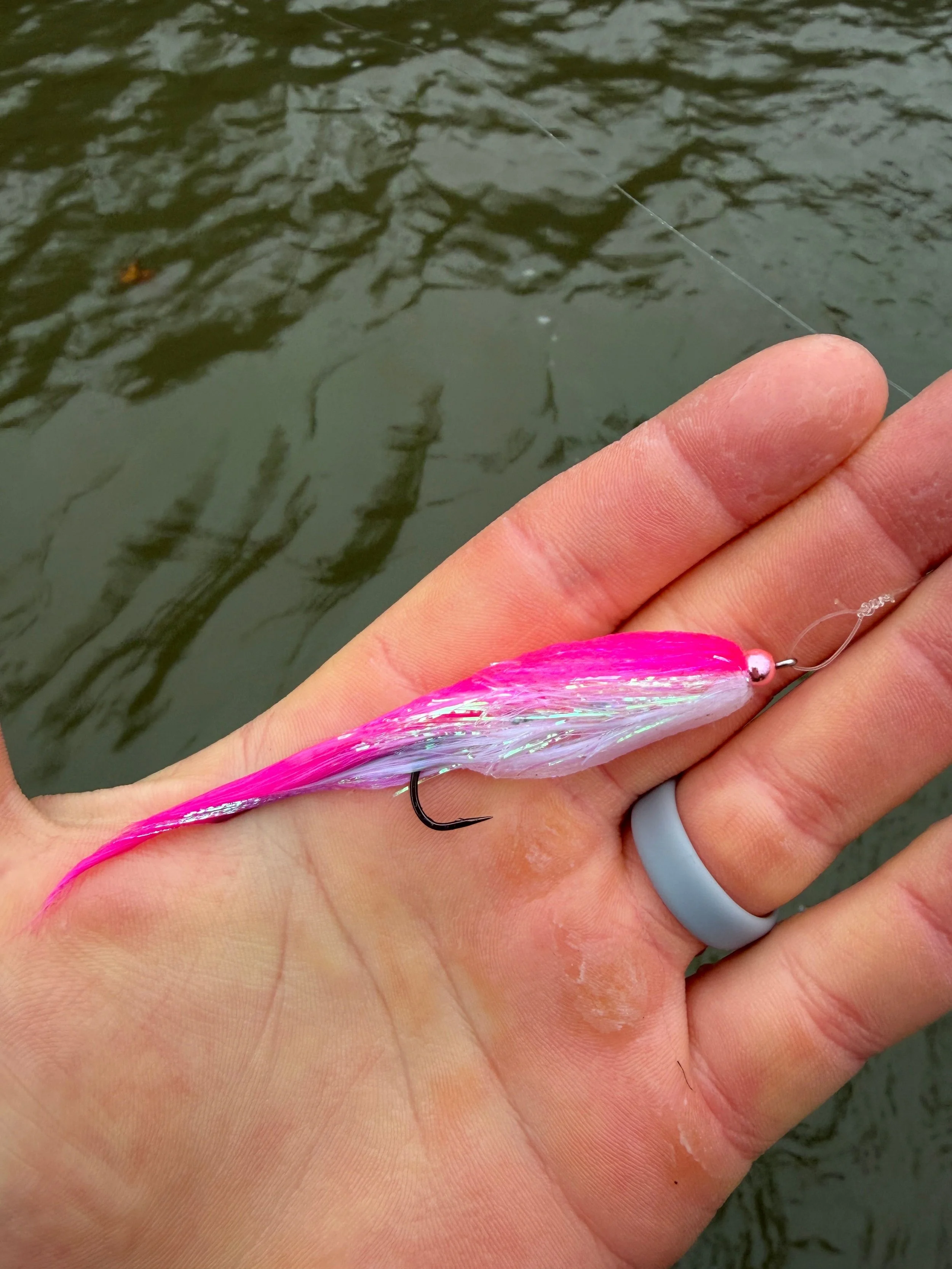 A person holding a pink and white fishing lure with a hook, over a body of water.