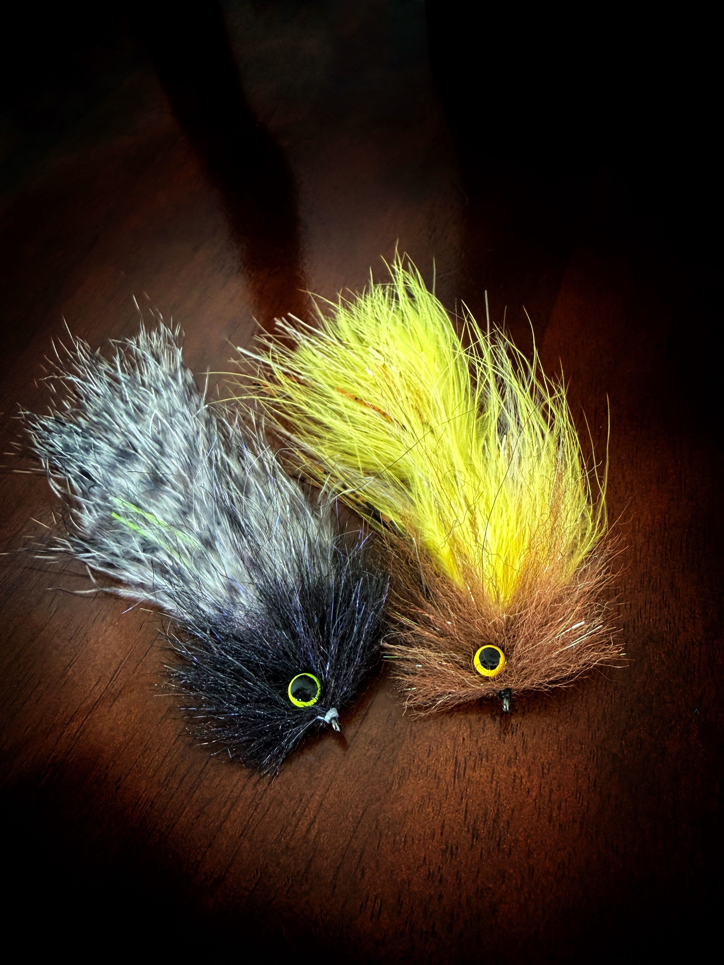 Two fishing lures designed to look like small furry animals with prominent yellow eyes, one with gray and black fur and the other with brown and yellow fur, placed on a wooden surface.