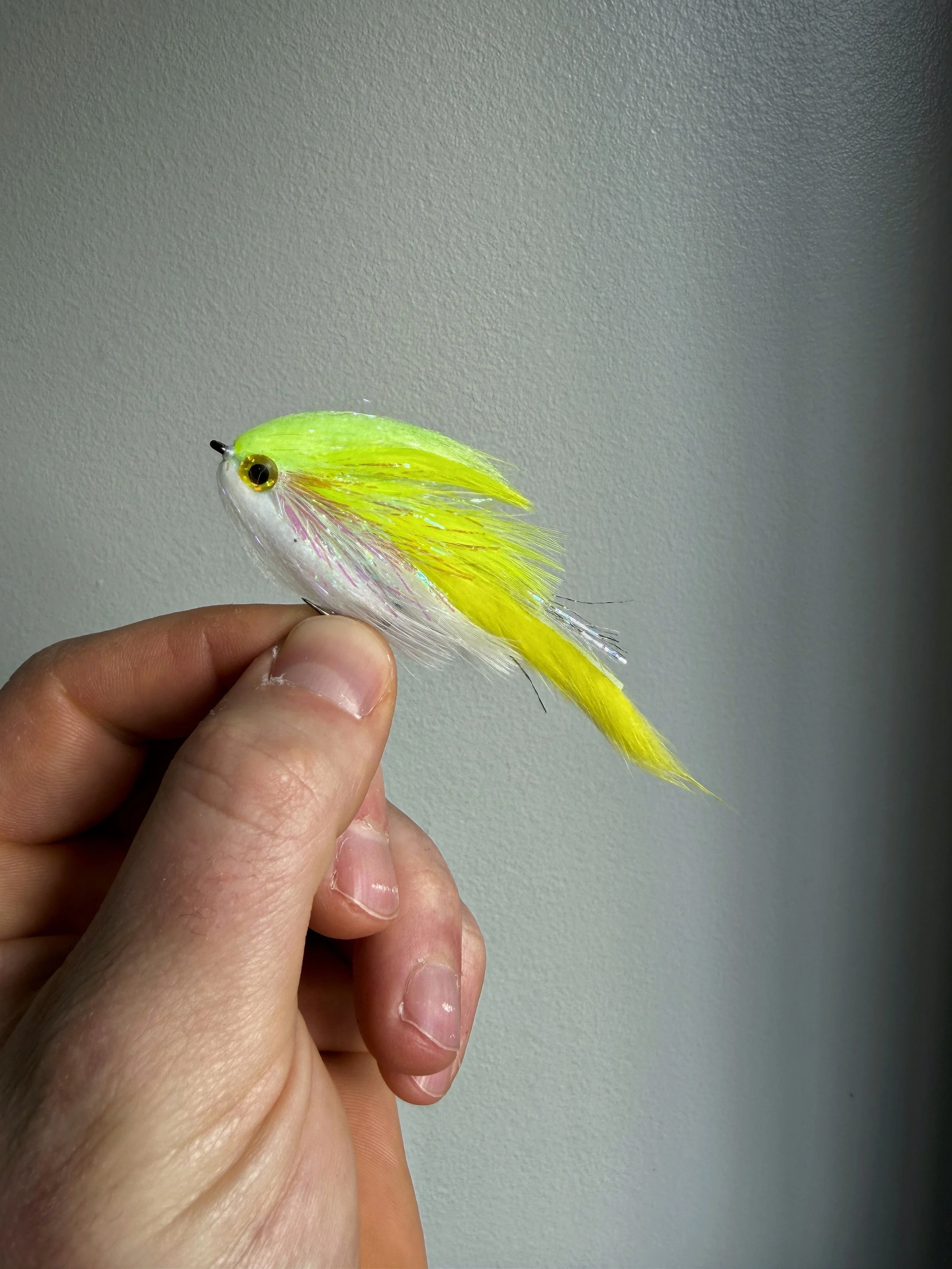 Hand holding a neon yellow and white fishing lure with feathers and a fish-shaped body against a plain wall background.