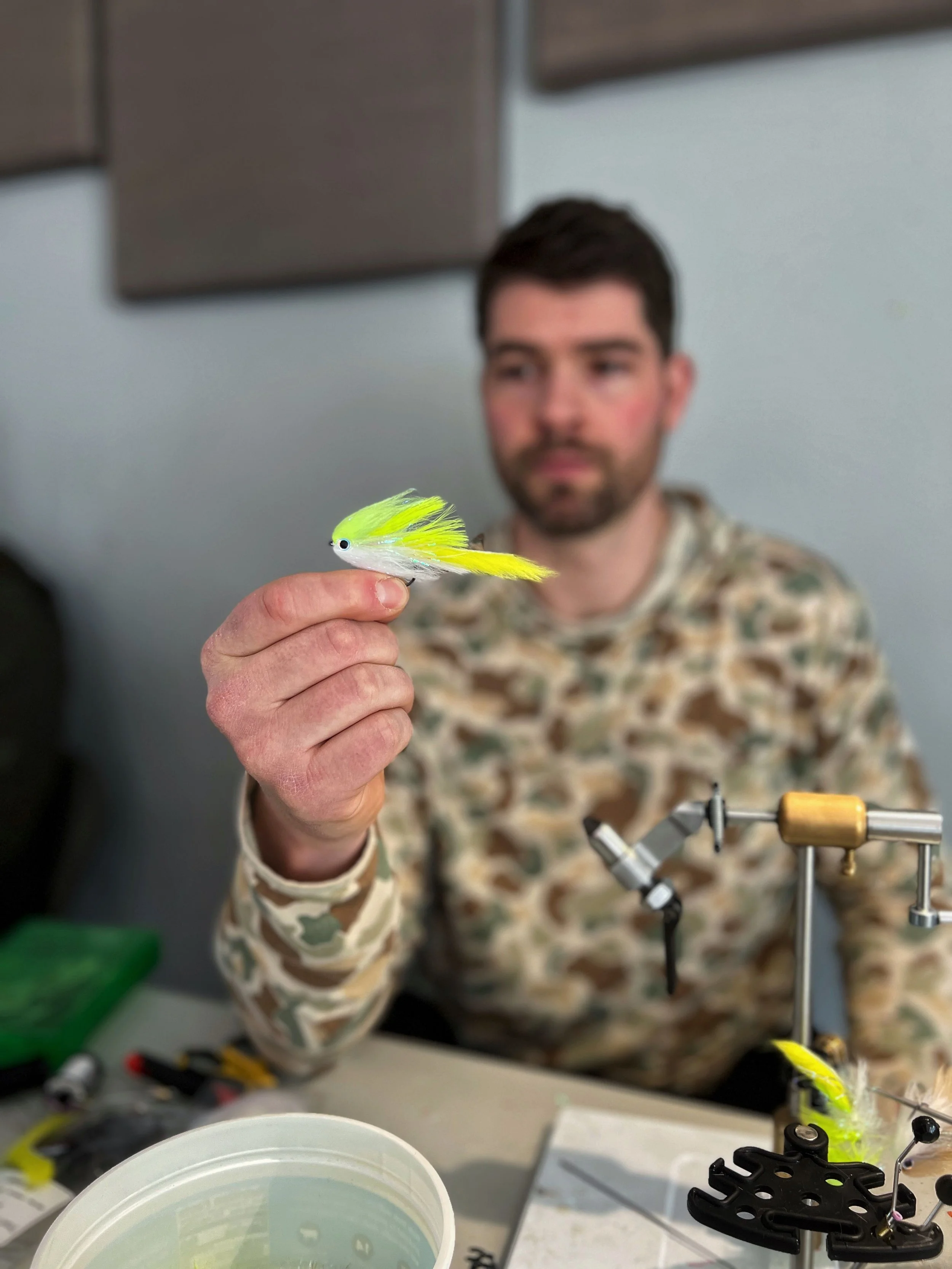 Man holding a yellow and white fishing lure in a workshop or fly-tying area, with fly-tying equipment and materials on the table.