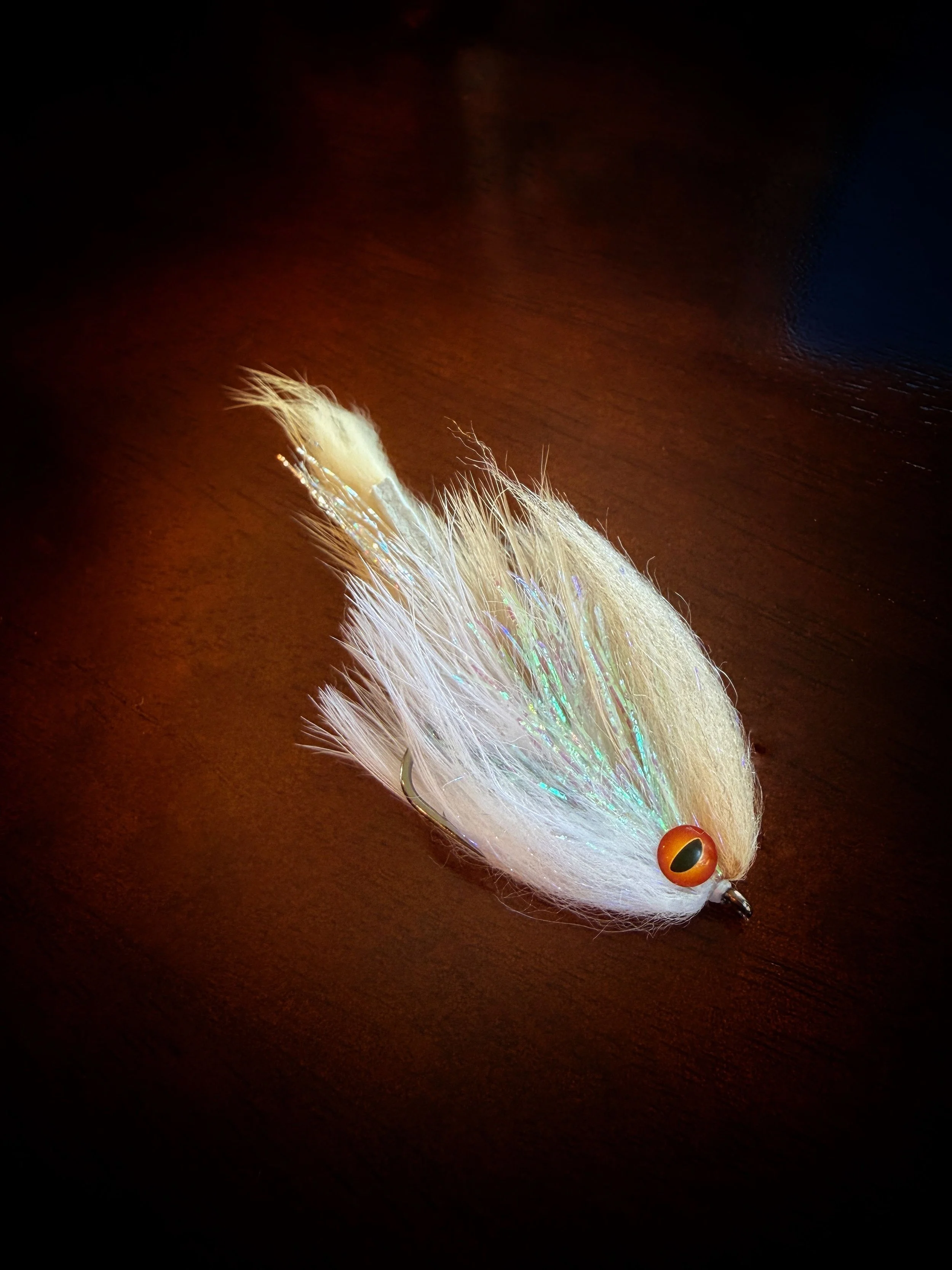 Close-up of a fishing lure with white and iridescent feathers and a large orange eye, resting on a dark wooden surface.