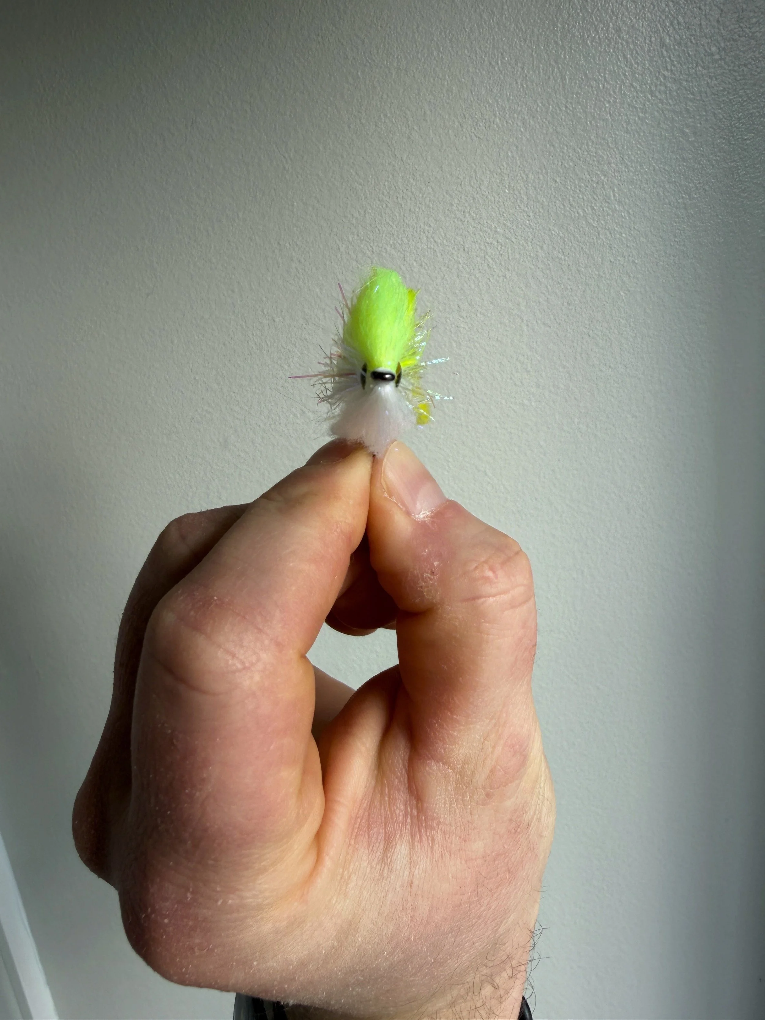 A person's hand holding a small fuzzy yellow and white fishing lure with a black eye, standing against a plain, light-colored wall.