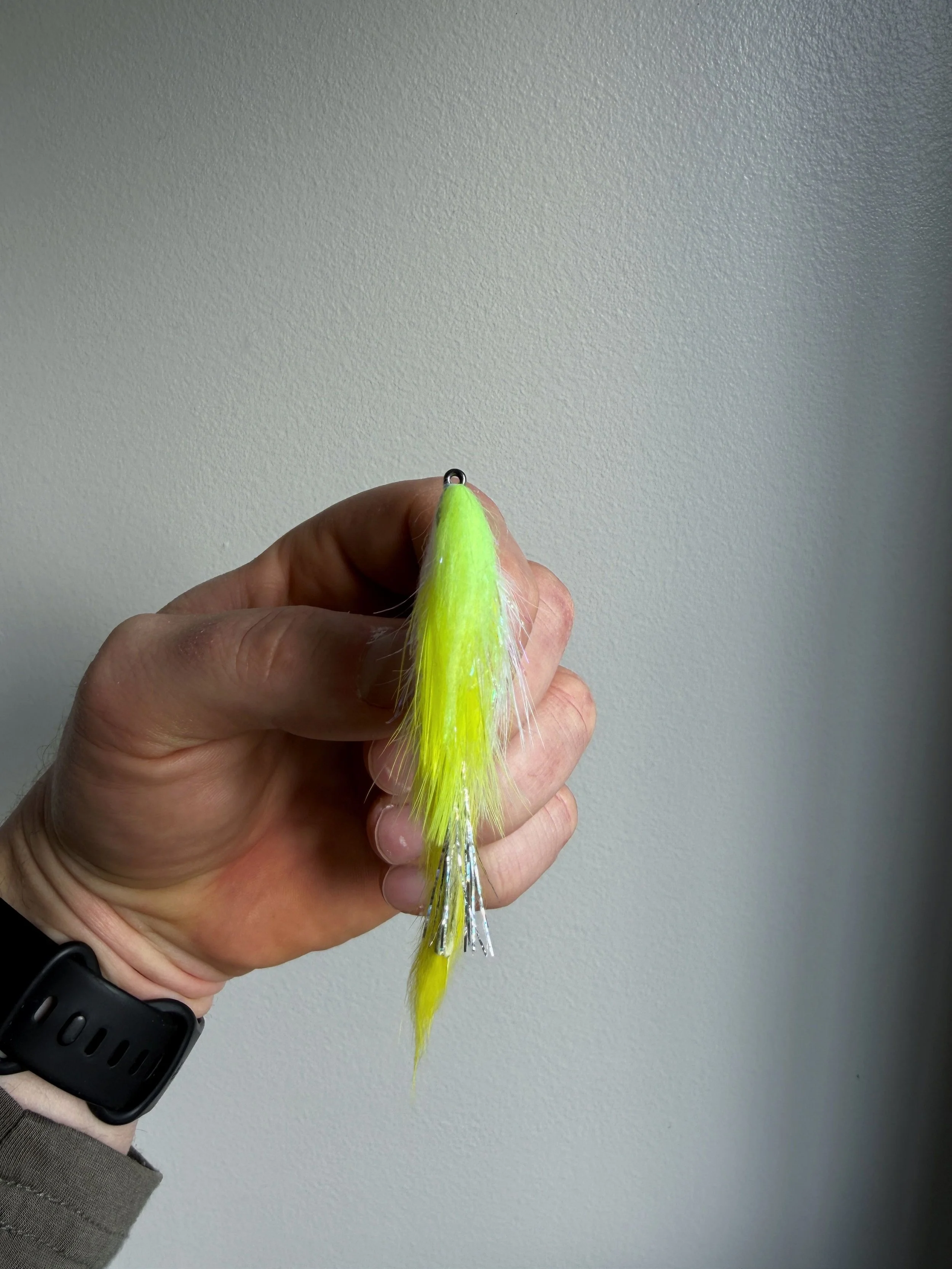 Person holding a fishing lure with bright yellow feathers and silver accents against a plain white wall background.