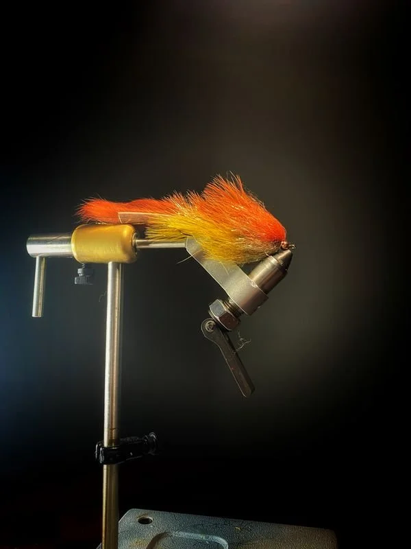 A close-up of a fishing fly with orange and yellow fur, mounted on a fly-tying vice against a black background.
