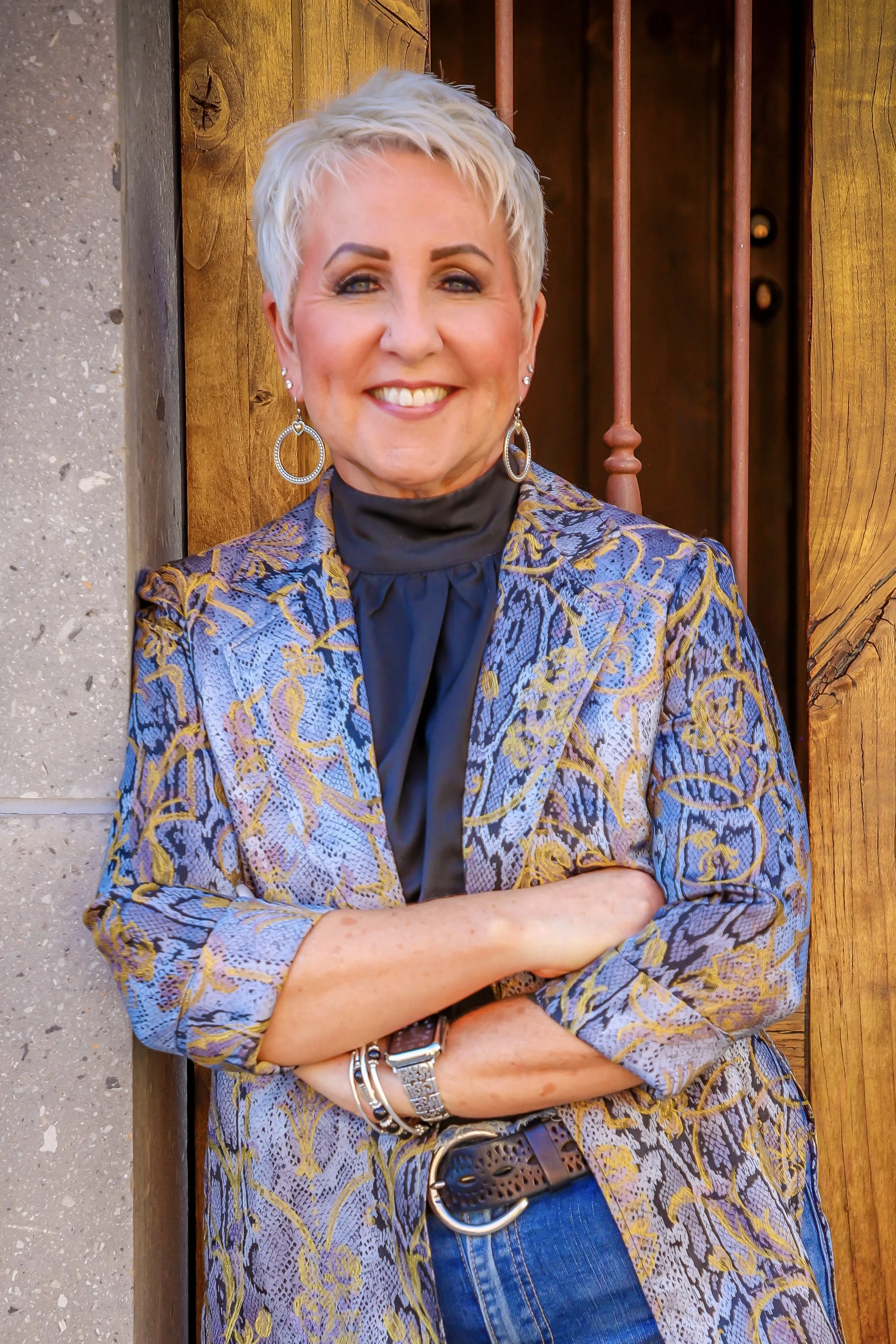 A woman with short platinum blonde hair, blue eyes, and makeup, wearing a patterned blazer with purple, gold, and black, a black top, blue jeans, and jewelry, standing with arms crossed in front of a textured concrete wall and wooden structure with metal bars.