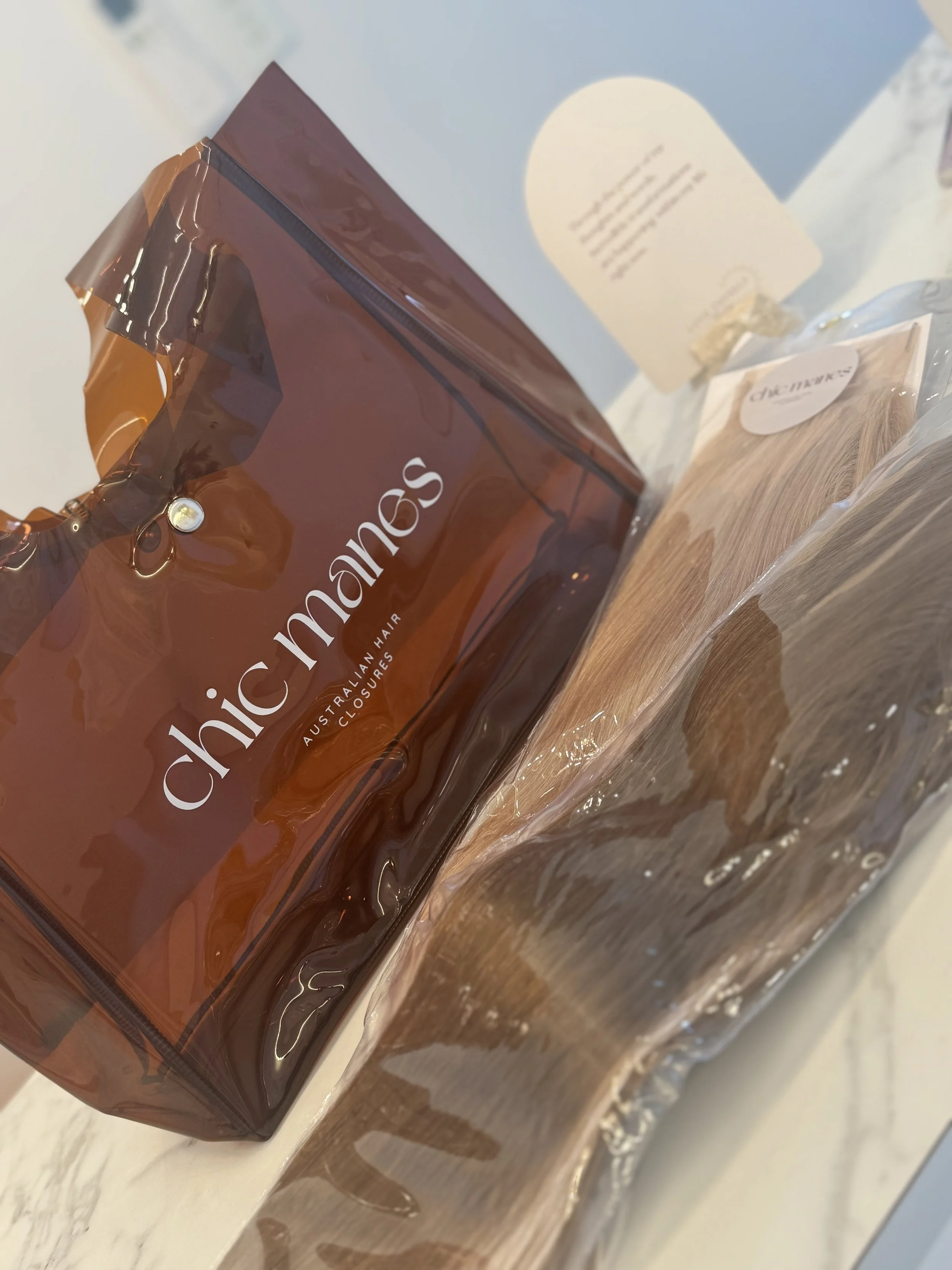 A brown bag of Australian hair closures labeled 'chic mane' on a white surface, with a wooden brush and some papers in the background.