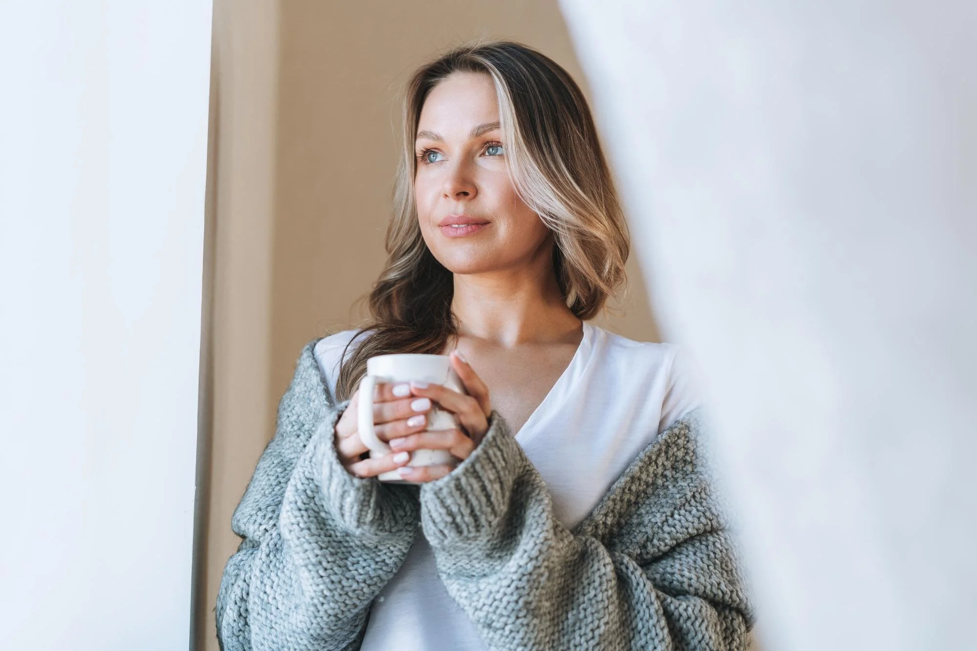 A woman with shoulder-length brown and blonde hair, blue eyes, and fair skin is holding a white mug with both hands. She is wearing a white top and a cozy gray knitted cardigan, standing indoors near a plain white wall, gazing thoughtfully.