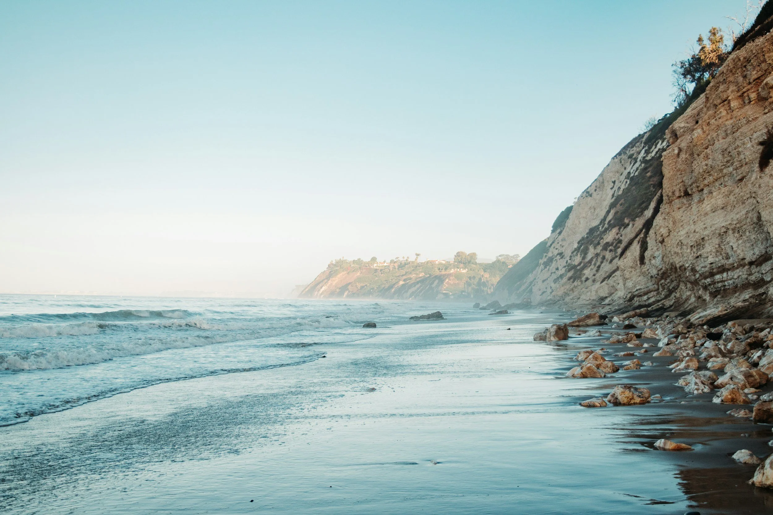 Coastal view with ocean waves hitting the sandy shoreline, rocky beach, and steep cliffs in the distance under a clear blue sky.
