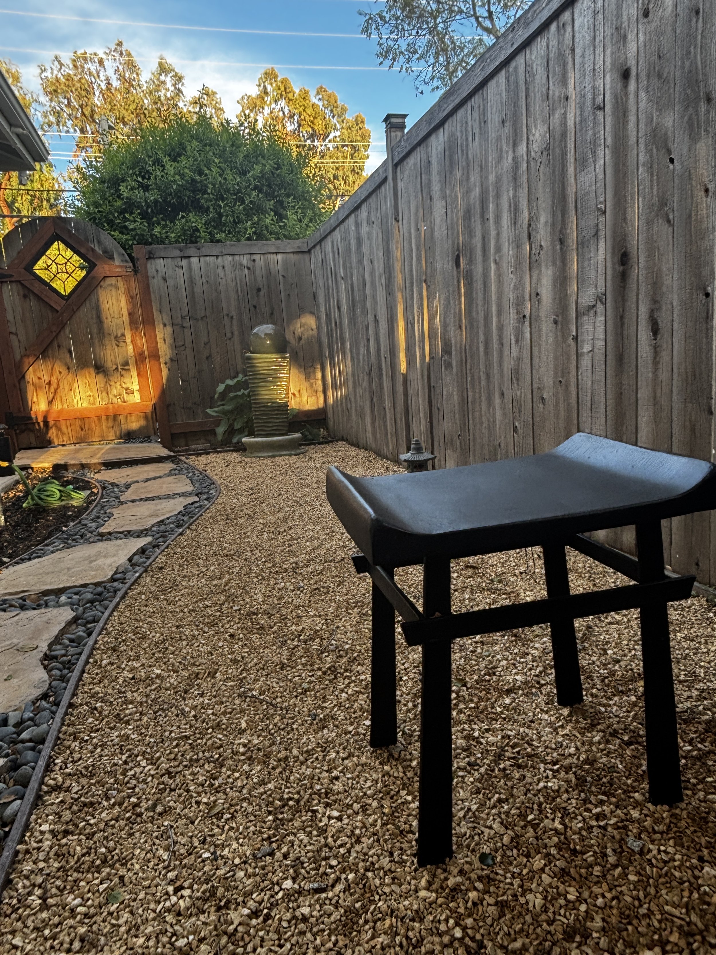 A backyard patio area with a gravel ground and a black metal table. There is a wooden fence with a gate in the corner, a water fountain, and a garden bed with stones and plants. Trees and a blue sky with clouds are visible in the background.