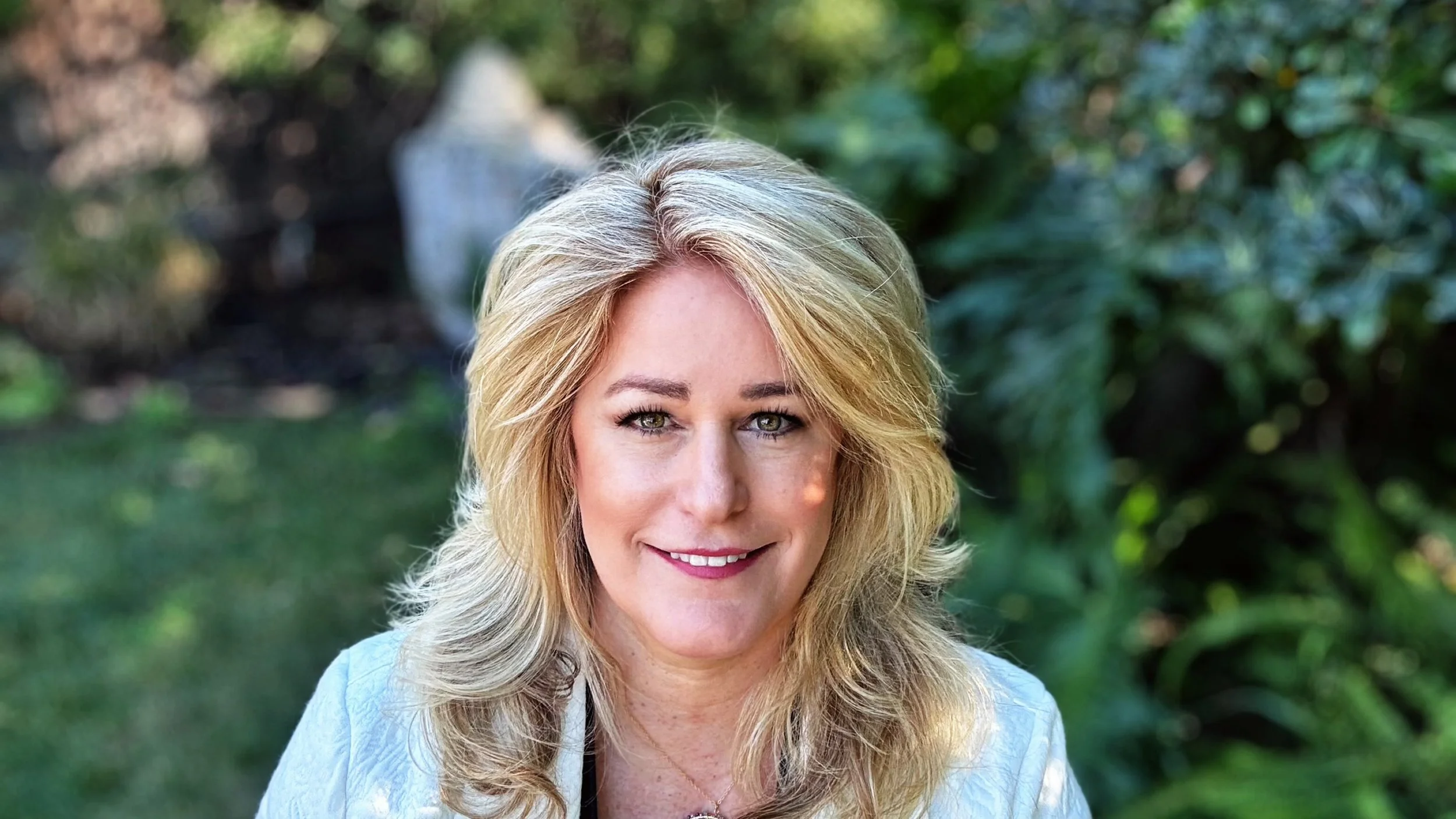 A portrait of a blonde woman with wavy hair, smiling and looking at the camera, outdoors with green foliage in the background.