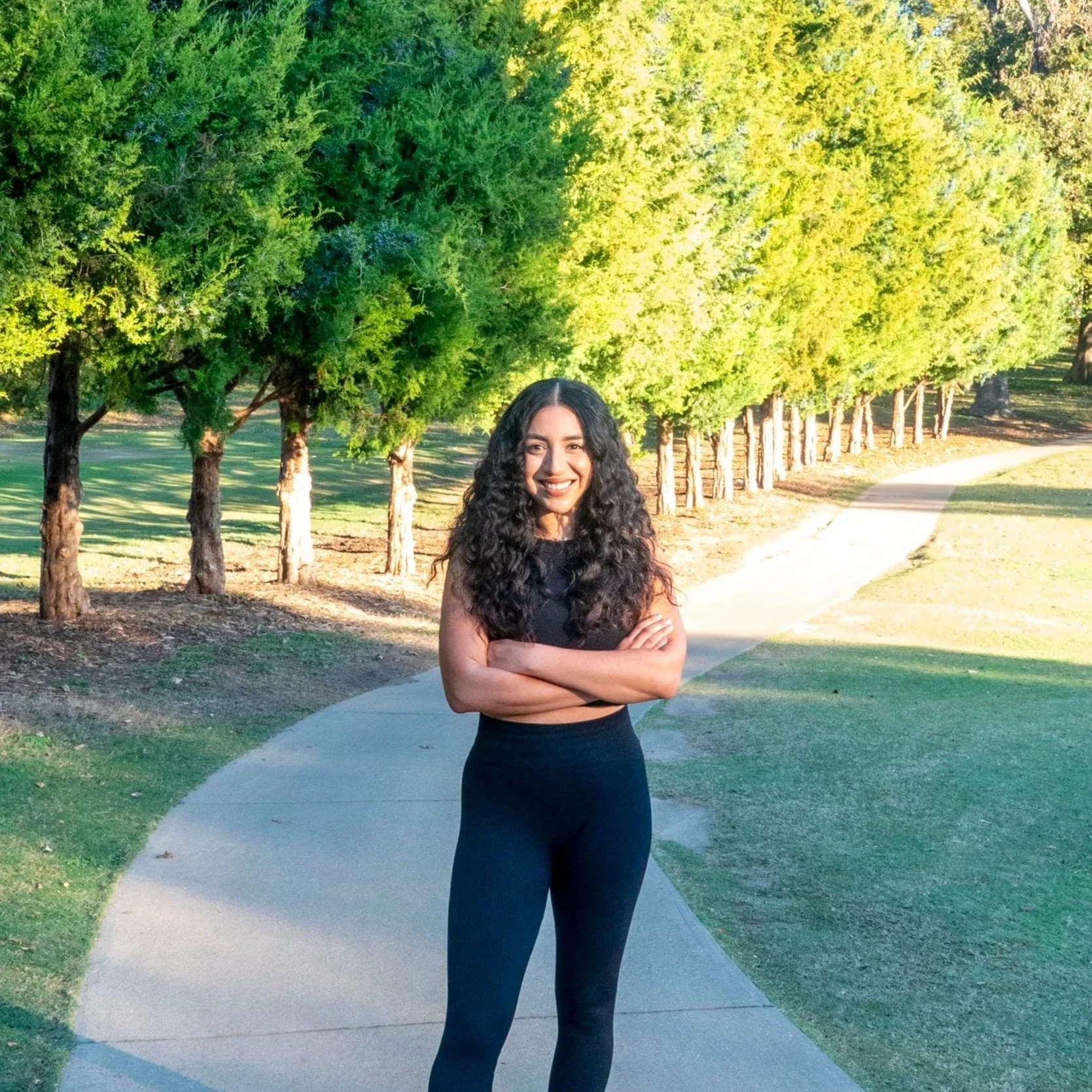 Young woman with long curly hair smiling and standing with crossed arms on a curved pathway in a park with green trees and grass on a sunny day.