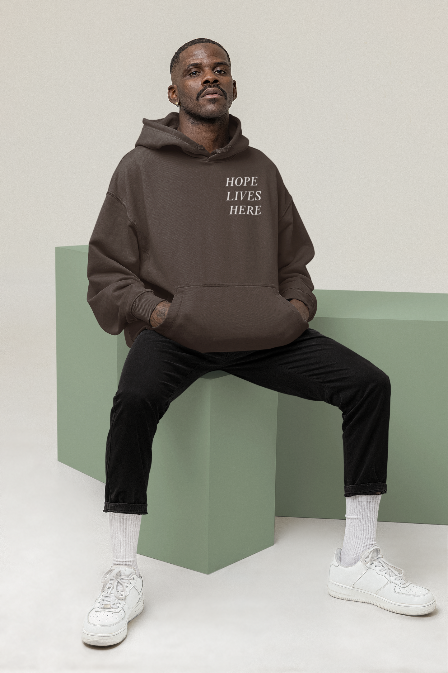 A man sitting on a green block, wearing a brown hoodie with the text 'HOPE LIVES HERE', black pants, white socks, and white sneakers, posing against a plain background.