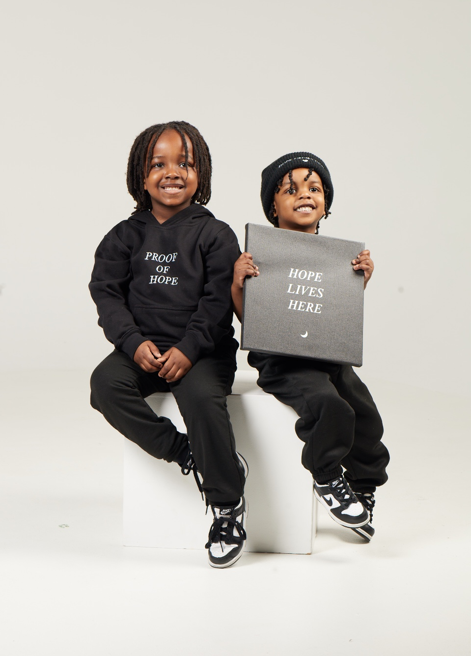 Two young children sitting on a white box against a plain background, one holding a sign that says "Hope lives here," both smiling and wearing black clothing.