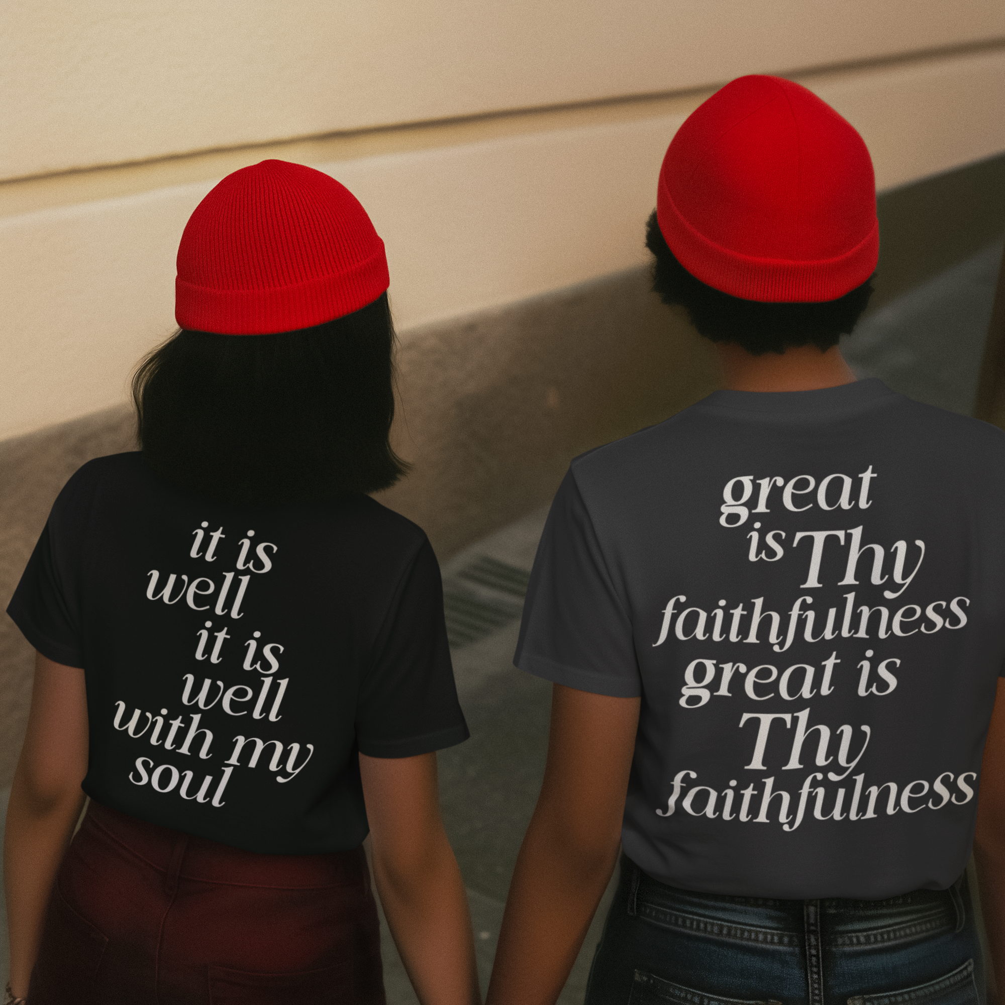 Two individuals wearing black and gray shirts with white text, each wearing a red beanie, walking with their backs to the camera.