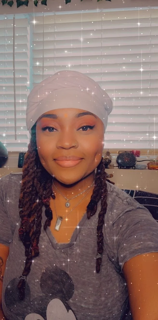 A woman with dreadlocks wearing a white head wrap, gray T-shirt with Mickey Mouse print, and layered necklaces, smiling at the camera. The background shows closed window blinds with decorative items on the window sill, and sparkles are overlaid on the photo.
