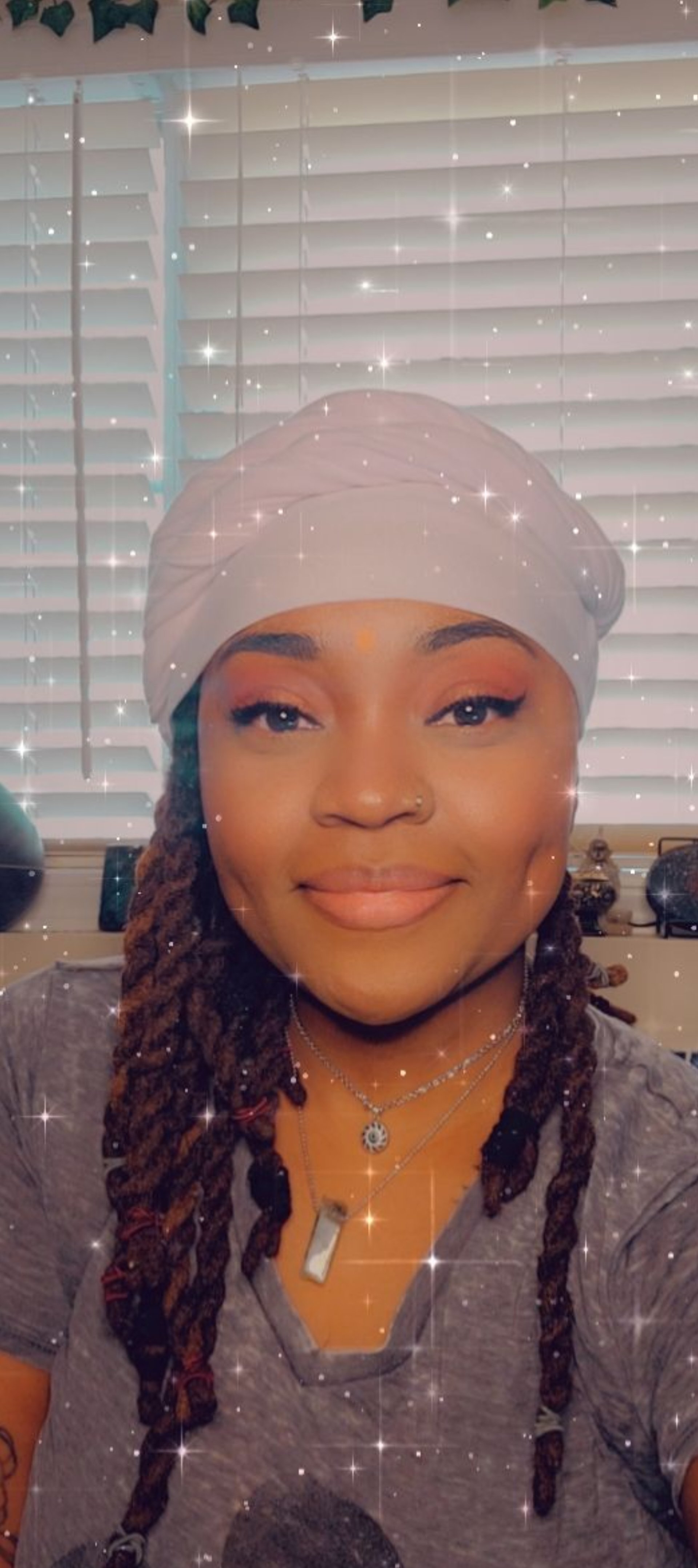 A woman with long braided hair, wearing a white headwrap, smiling at the camera with a sparkly filter effect, in front of window blinds.