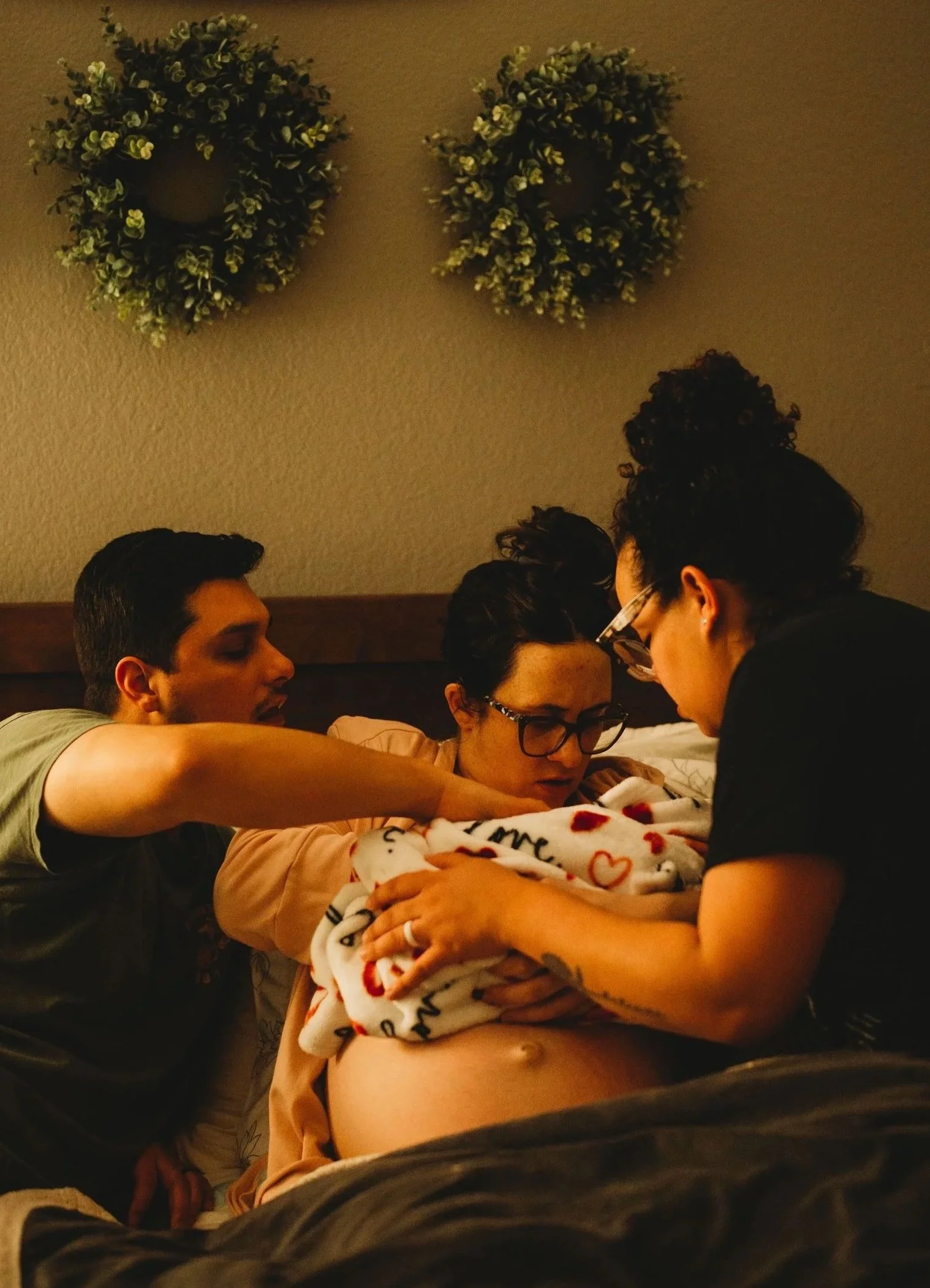 A woman unpacks her newborn baby with support from her spanish speaking doula in a bedroom, with two decorative wreaths on the wall. In Austin, Texas.