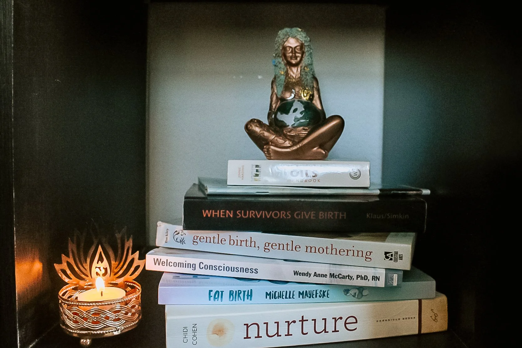 Stack of books on a shelf with a small burning candle to the left, and a brass statue of a seated woman with curly hair holding a globe on top of the books.