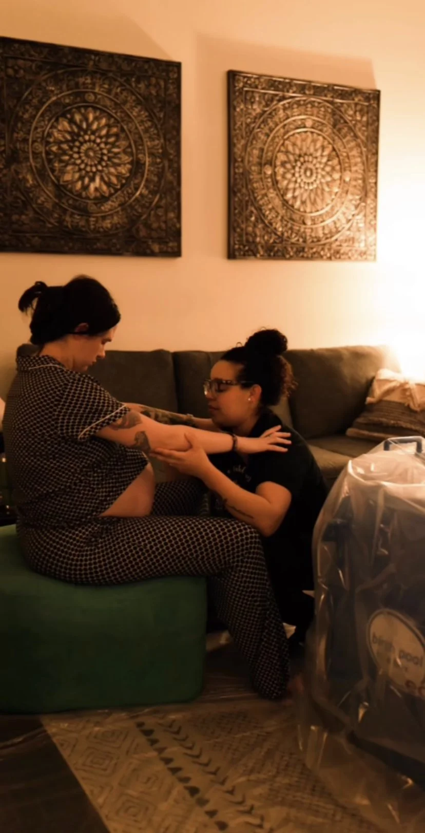 A woman laboring while bending over on a bed, receiving a traditional rebozo techniques from her Spanish speaking doula standing beside the bed in a cozy room with white curtains and a chest of drawers. In Austin, Texas.