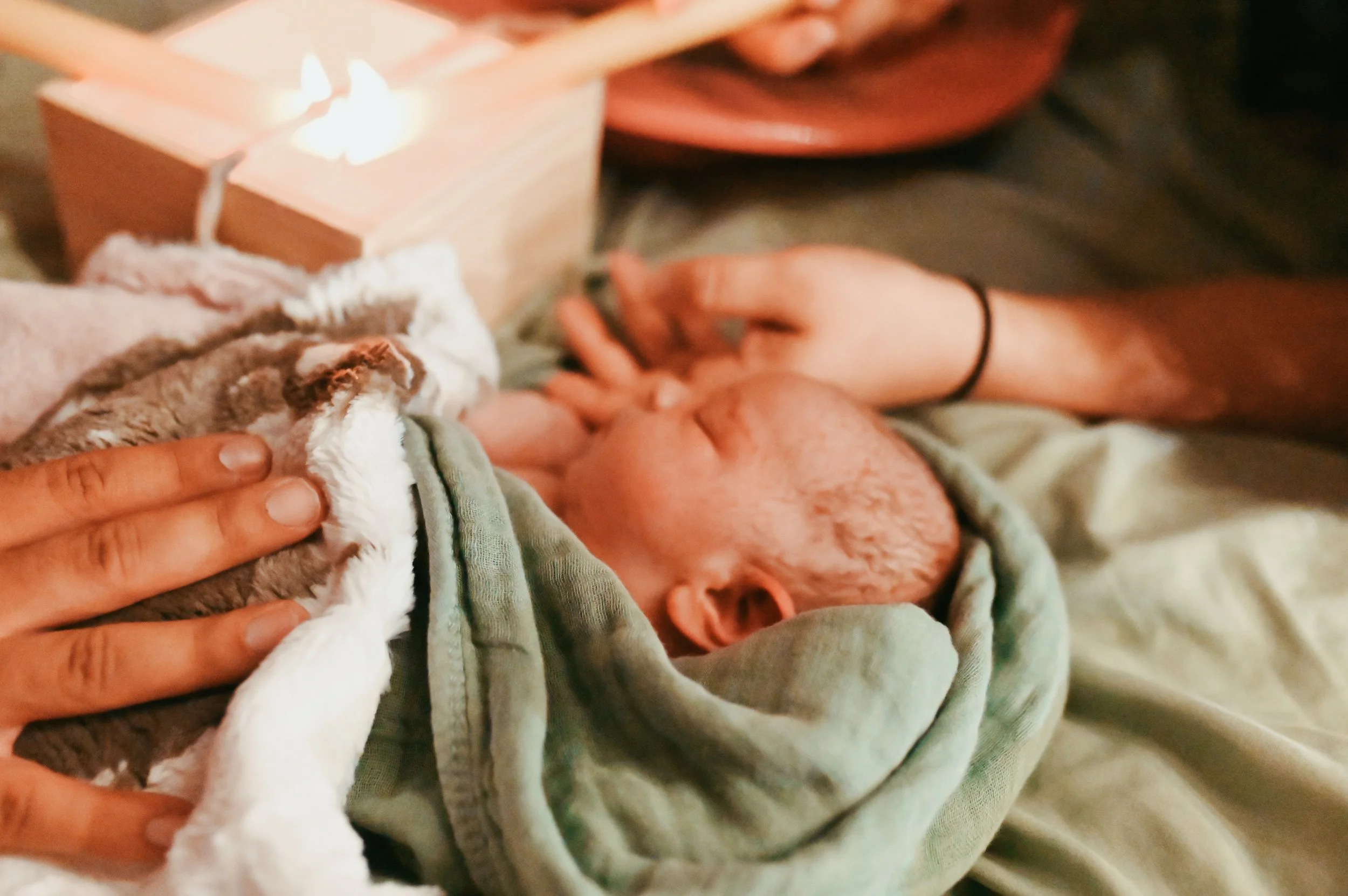 A spanish speaking doula is holding a newborn baby wrapped in a blanket, with the baby's head resting on her arm. There is a lit candle in a wooden holder on a nearby table, creating a warm, intimate atmosphere as they burn the umbilical cord.