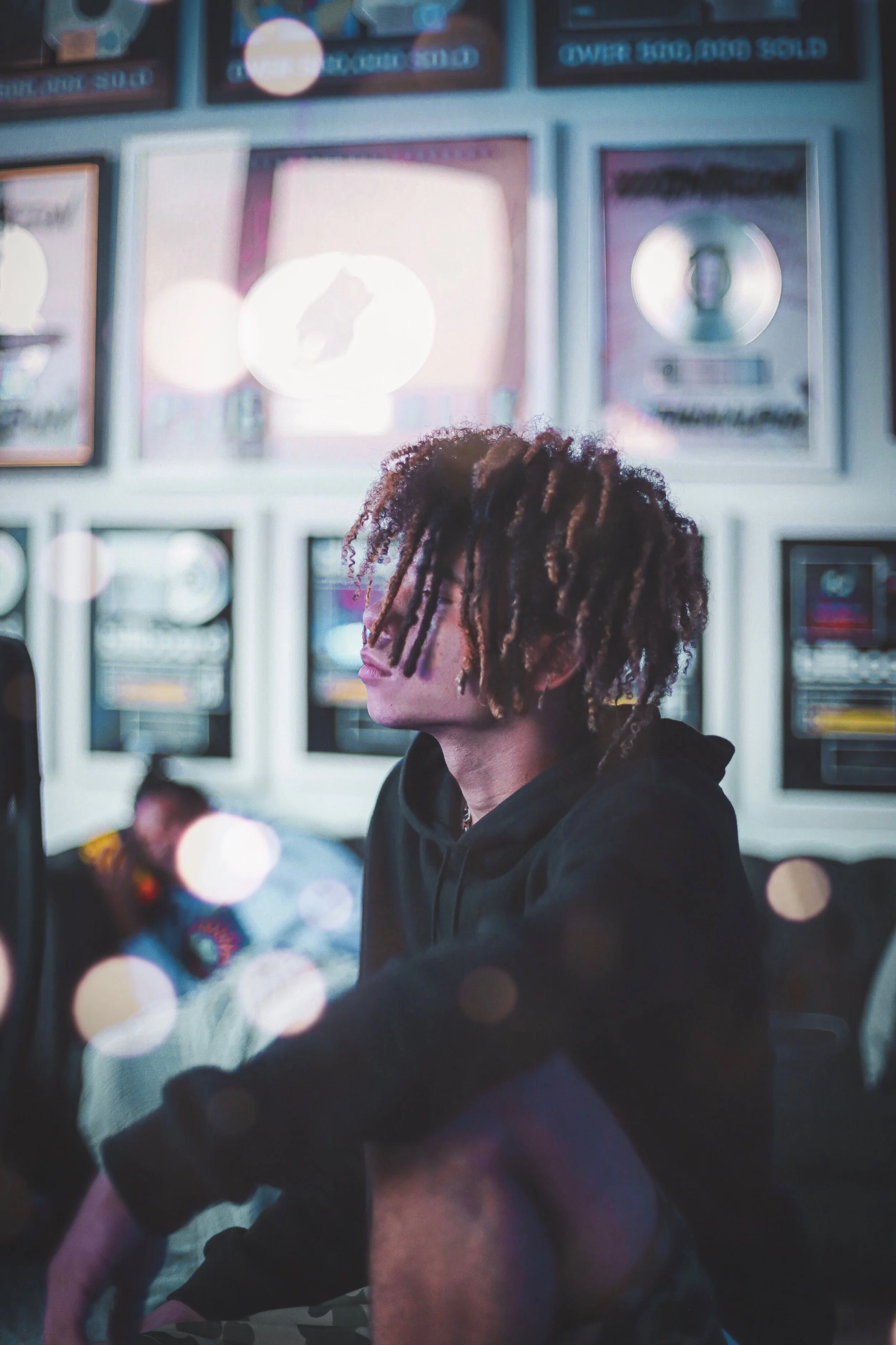 A young man with dreadlocks sitting in an arcade, surrounded by framed video game awards and posters, with bright lights and a bokeh effect.