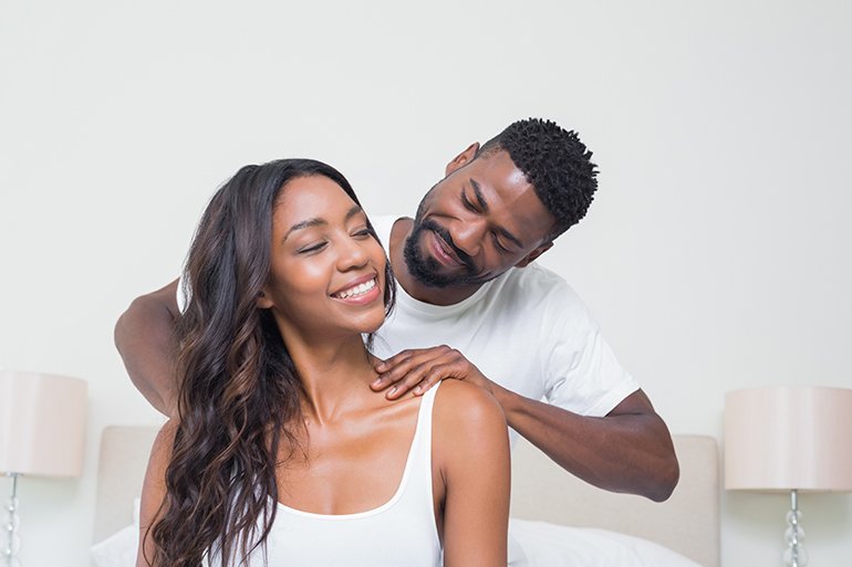 A man giving a shoulder massage to a woman after couples massage class, in a bedroom, both smiling and looking relaxed.