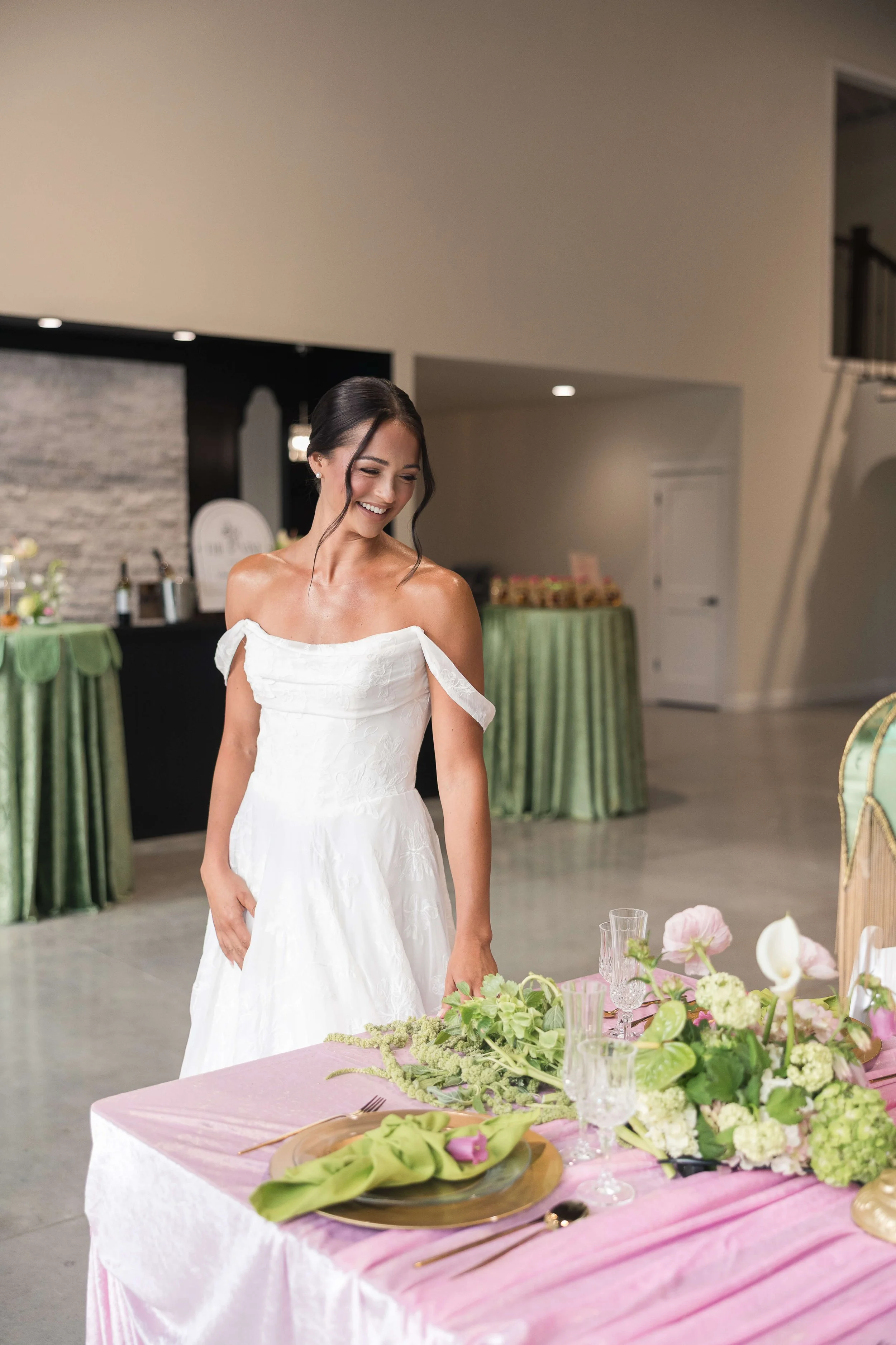 A woman in a white dress at a wedding reception, smiling and looking down, standing near a dining table decorated with pink and green floral arrangements, glassware, and table settings, in an indoor venue with beige walls and green covered tables in the background.