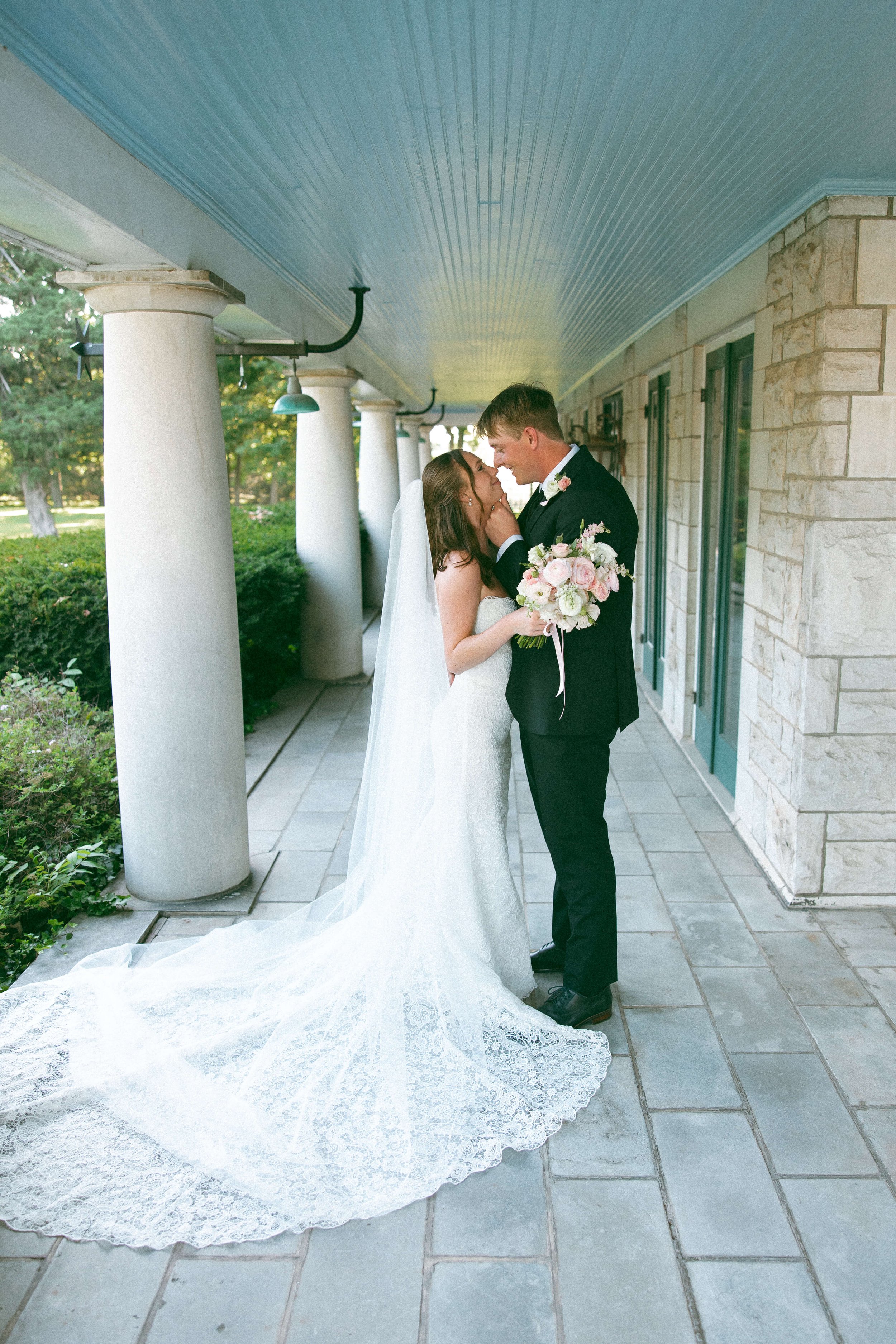 A bride and groom standing close together outside under a covered walkway, touching foreheads and smiling at each other. The bride holds a bouquet of pink and white flowers, and her long wedding dress flows onto the tiled floor.