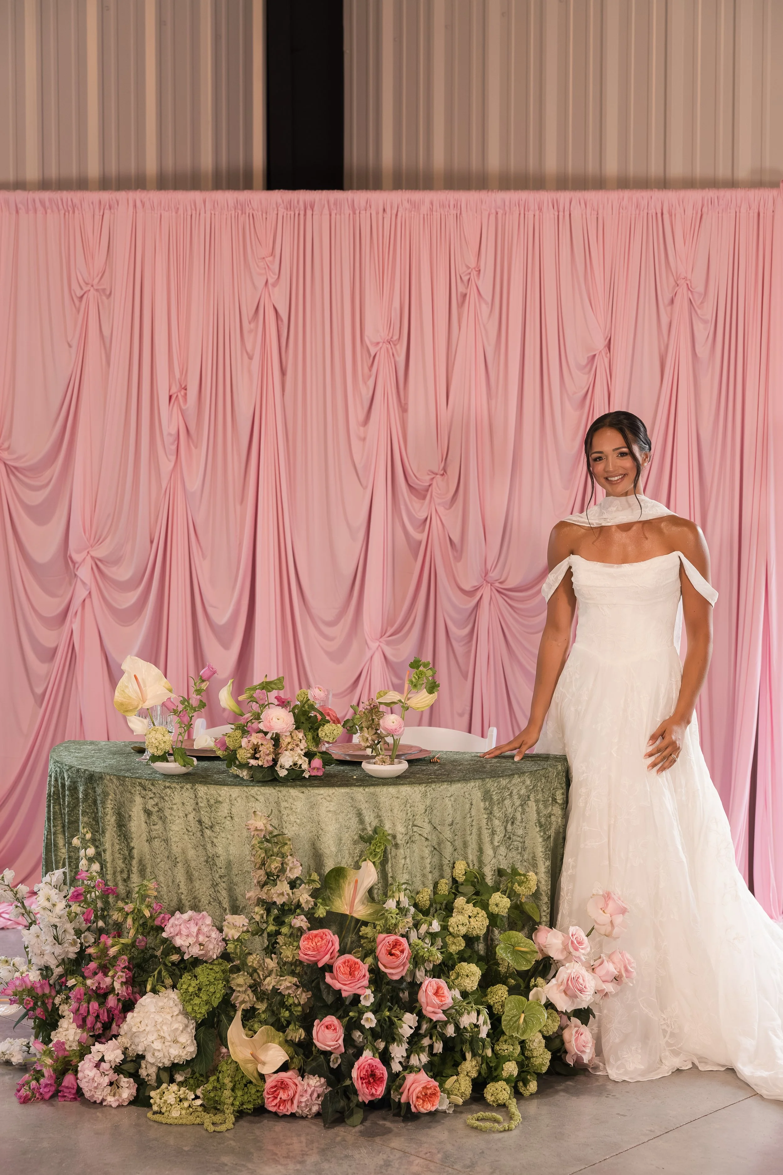 A woman in a wedding dress standing next to a table decorated with flowers and greenery, with a pink curtain backdrop. The table has a floral centerpiece and some dishes. There are many flowers on the floor in front of the table.
