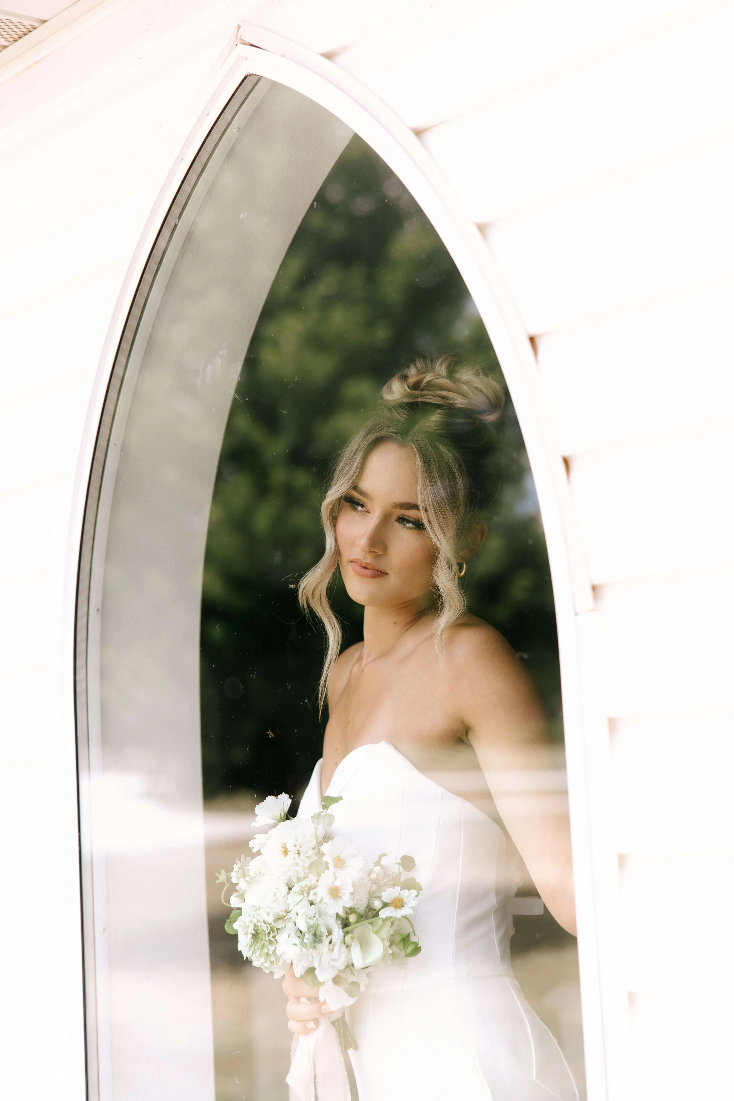 A bride looking out a window, holding a bouquet of white flowers, wearing a strapless white wedding dress, with her hair styled in a loose updo.