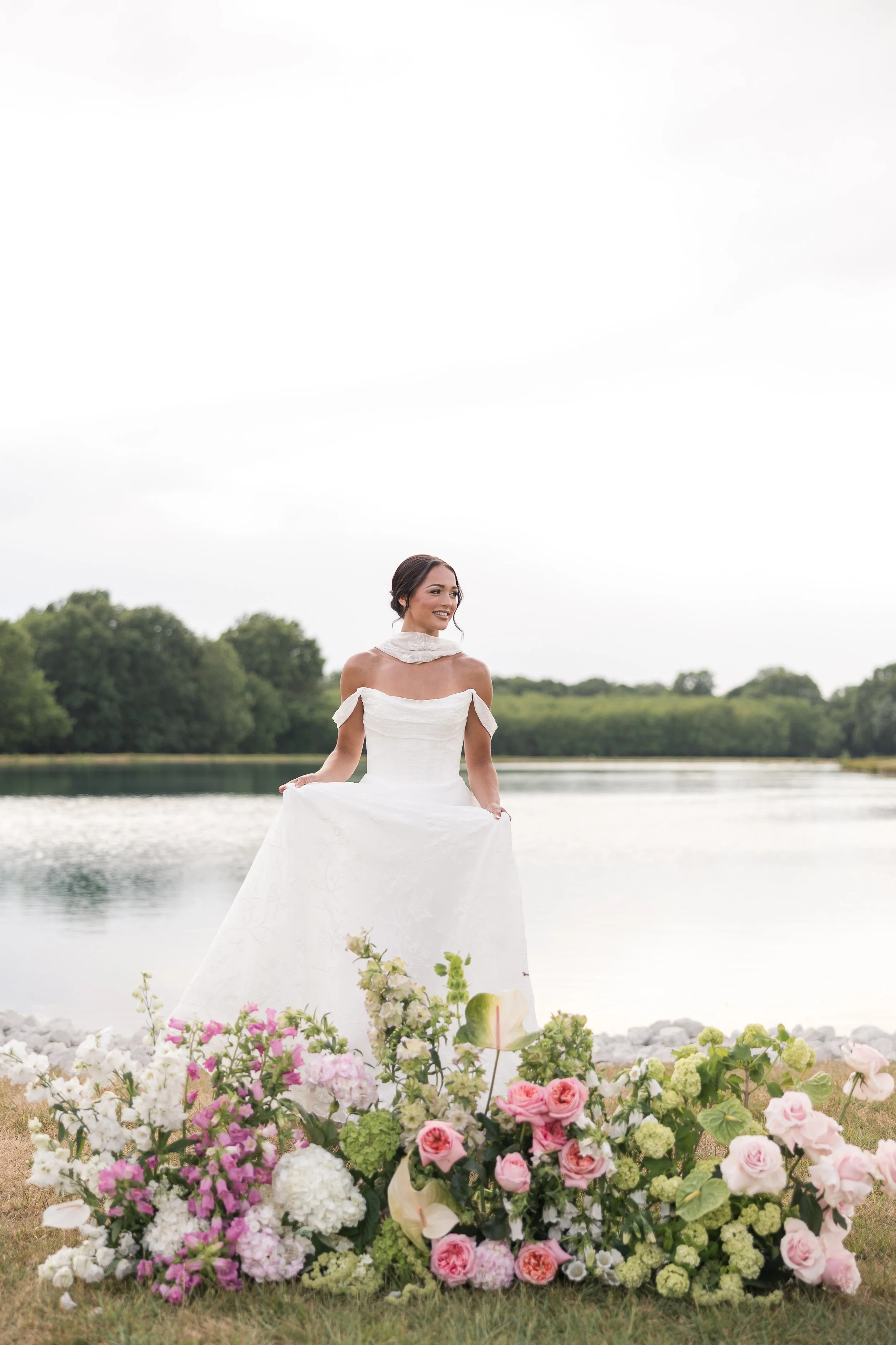 A bride in a white wedding dress holding her skirt, standing by a lake surrounded by pink, white, and cream flowers, with trees and overcast sky in the background.