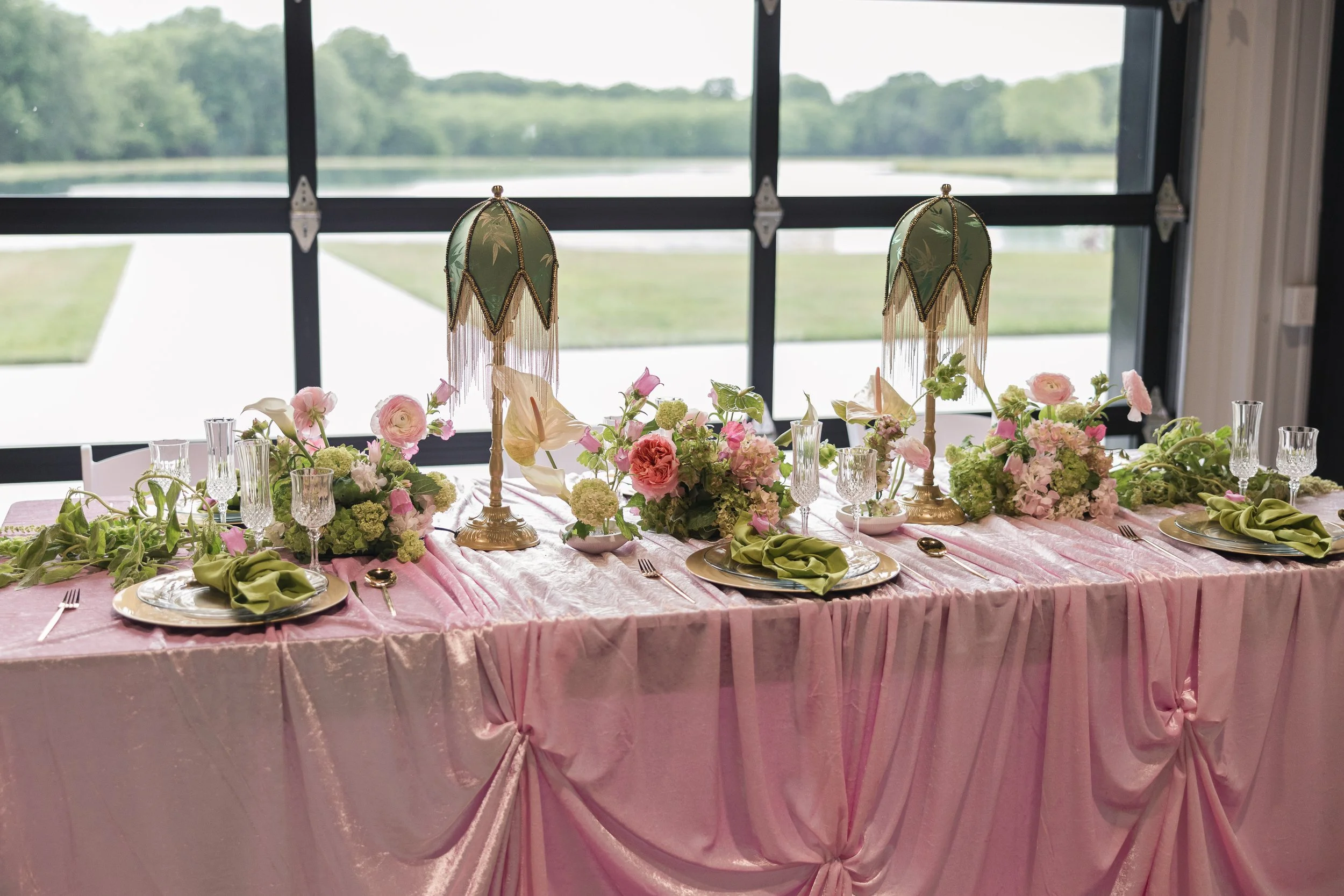 Decorated banquet table with pink tablecloth, floral centerpieces, gold and glass tableware, and green napkins, set in front of large window overlooking a grassy landscape and water.