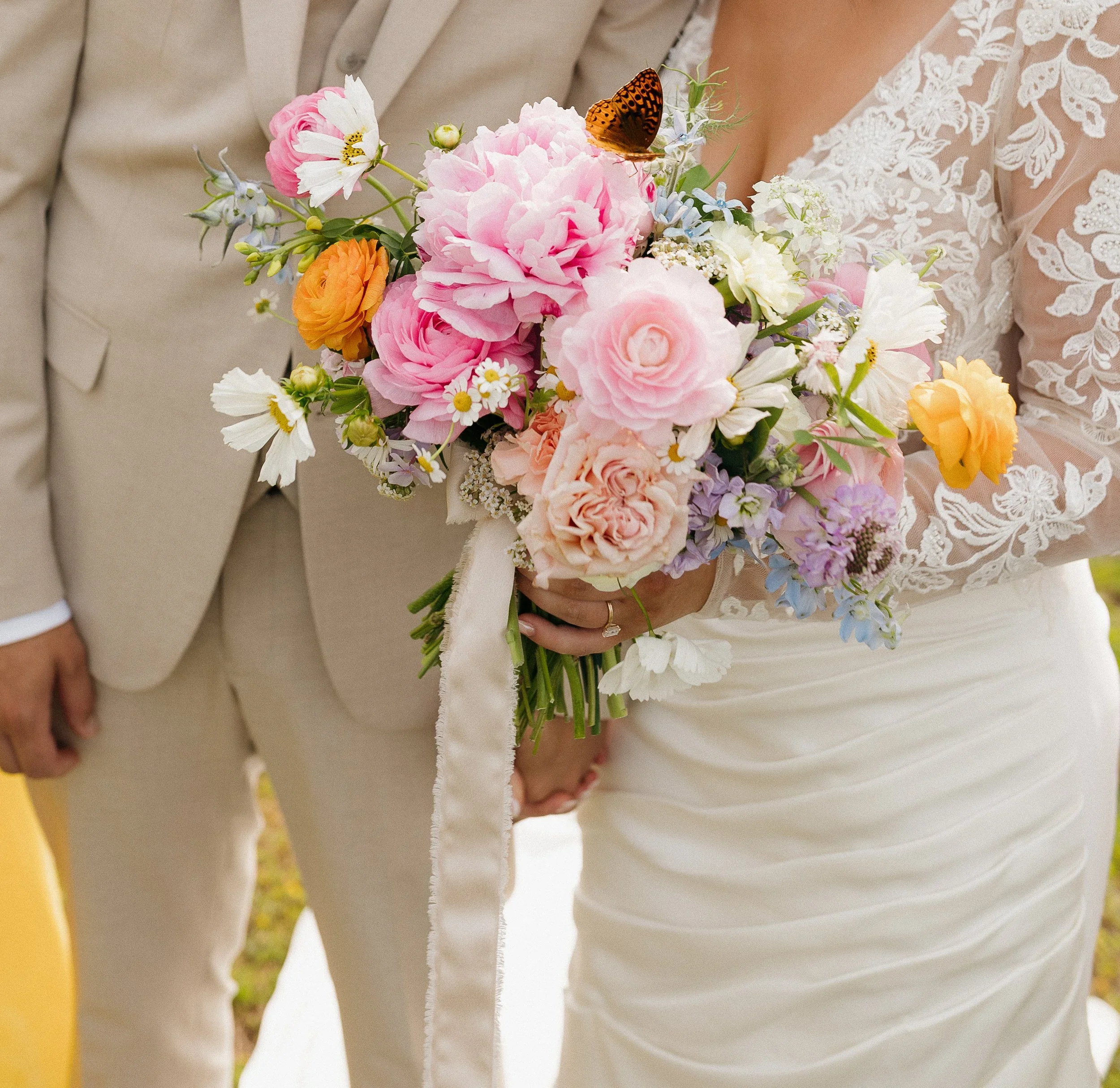 A bride holding a bouquet of pink, orange, white, and purple flowers, with a butterfly perched on the bouquet. The bride is wearing a white lace dress, and other people in beige suits are visible nearby, suggesting a wedding ceremony.