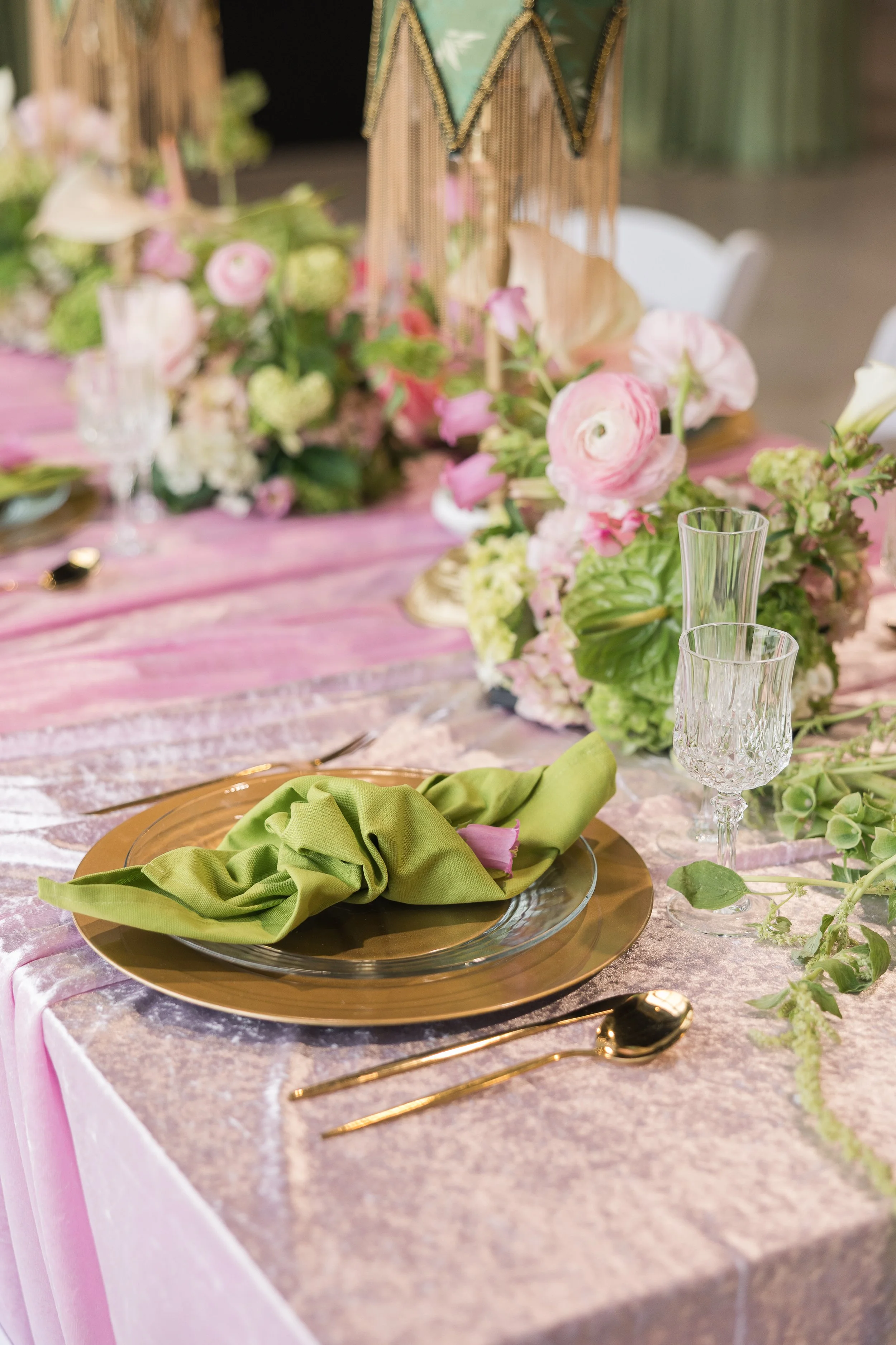 Elegant table setting with a green cloth napkin on a gold charger plate, clear glassware, and floral centerpiece with pink and white flowers on a pink and silver tablecloth.