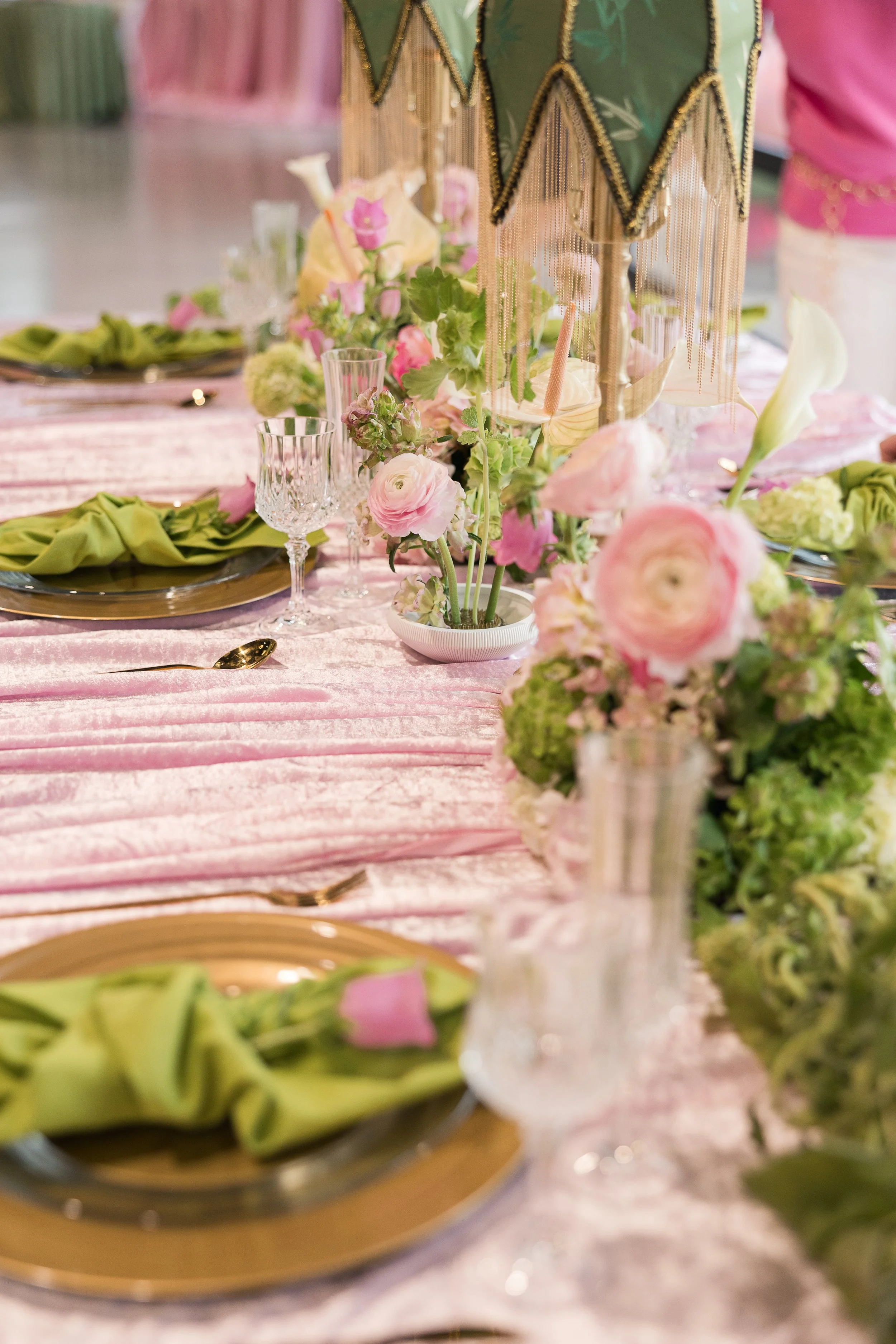 A decorated banquet table with pink tablecloth, floral centerpieces, crystal glassware, gold-rimmed plates, and green napkins.