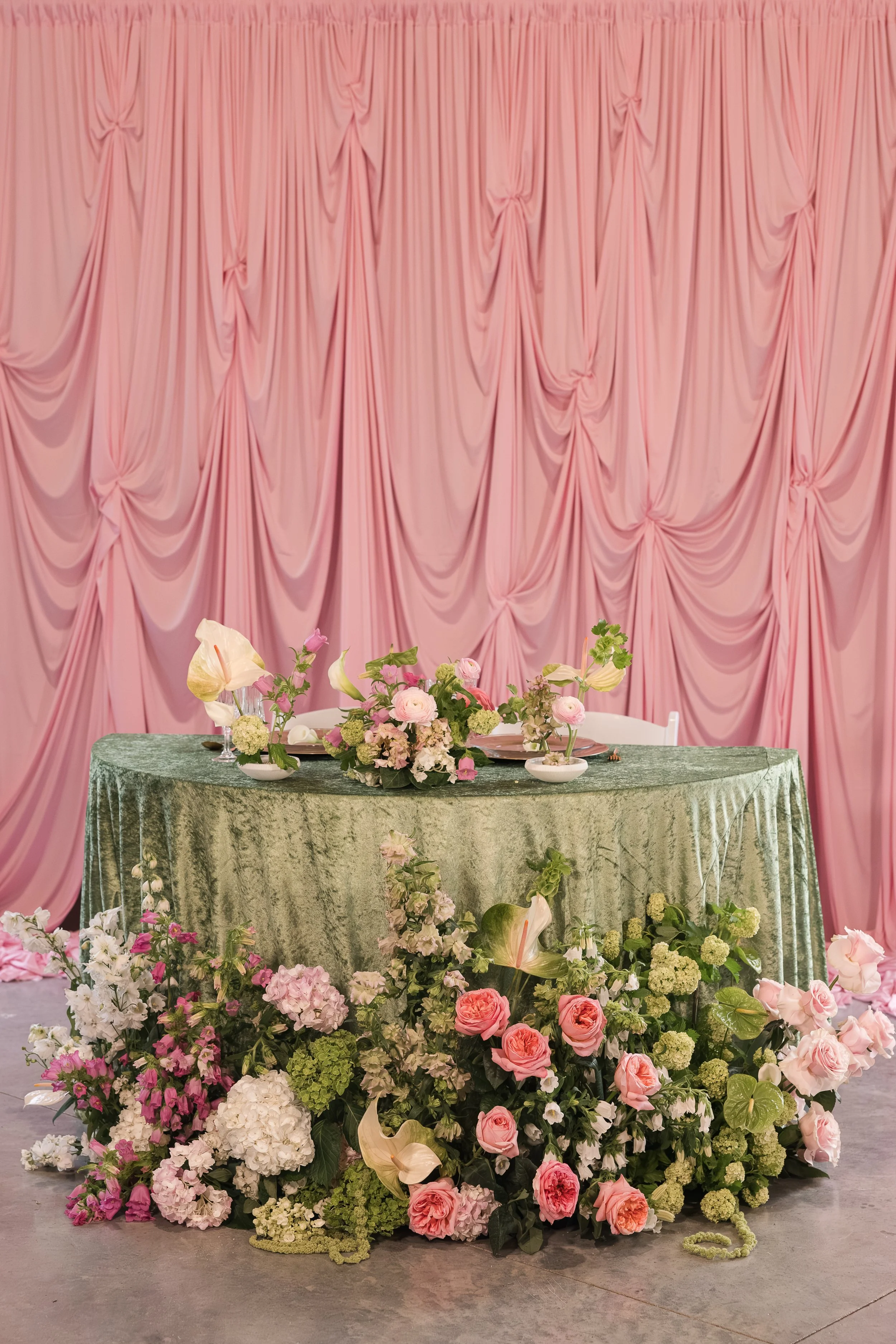 A decorated banquet table with an elaborate floral arrangement, set against a pink draped backdrop.