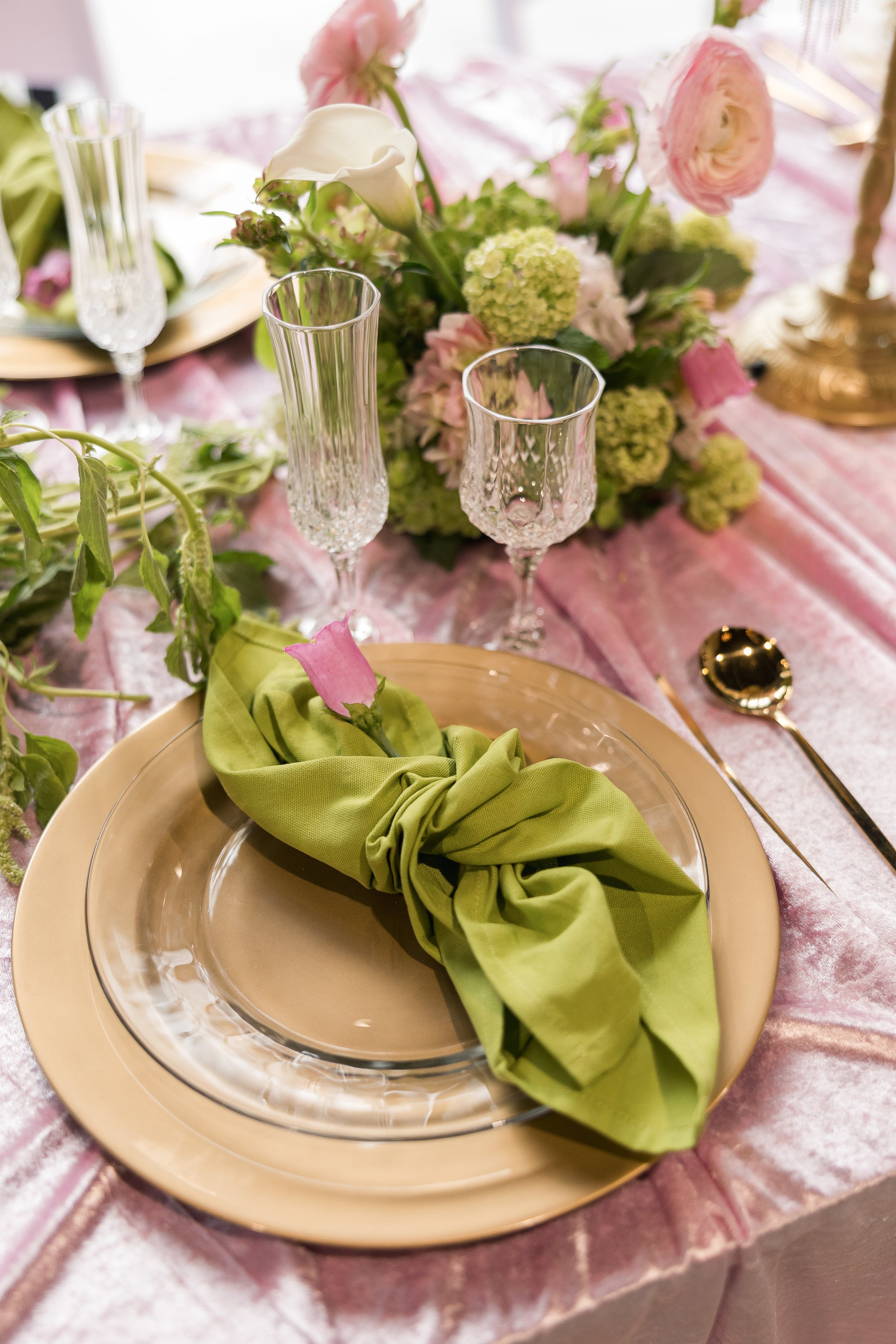 A formal table setting with a beige plate, a green cloth napkin, crystal glasses, and a floral centerpiece with pink and white flowers on a pink tablecloth.