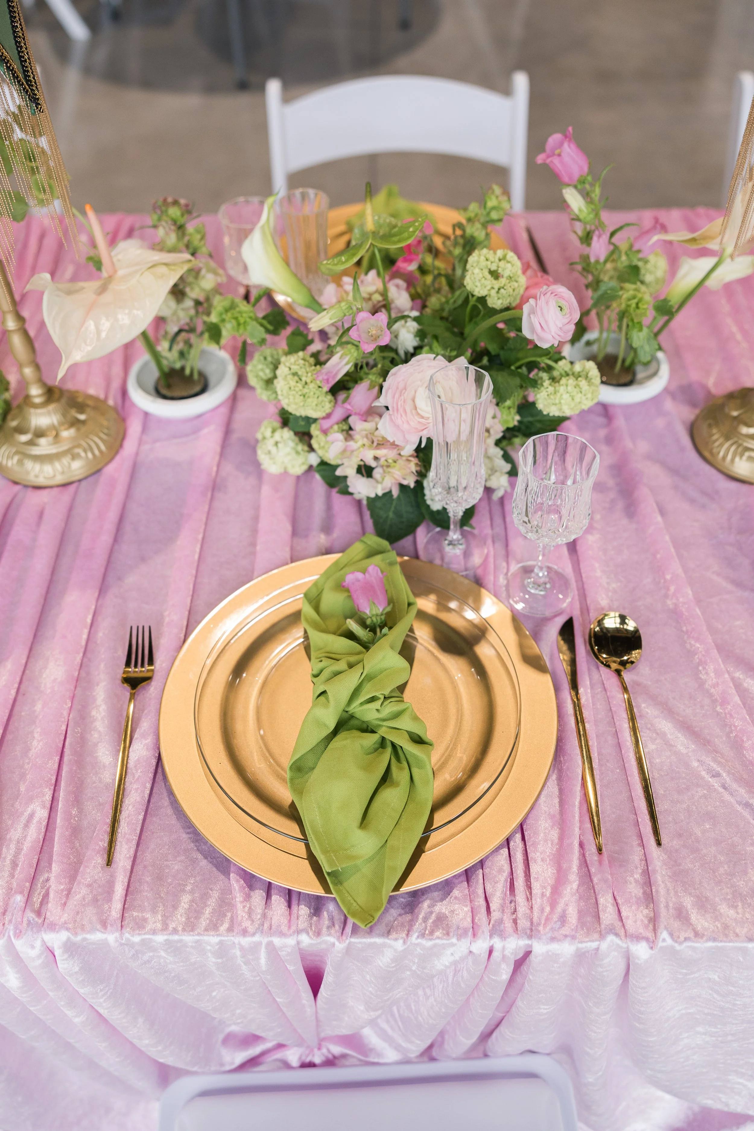 A pink tablecloth-draped table set for a formal event, featuring a gold charger plate with a green cloth napkin tied around a pink flower, gold flatware, clear glassware, and a floral centerpiece with pink and white flowers. Gold candlestick lamps ar