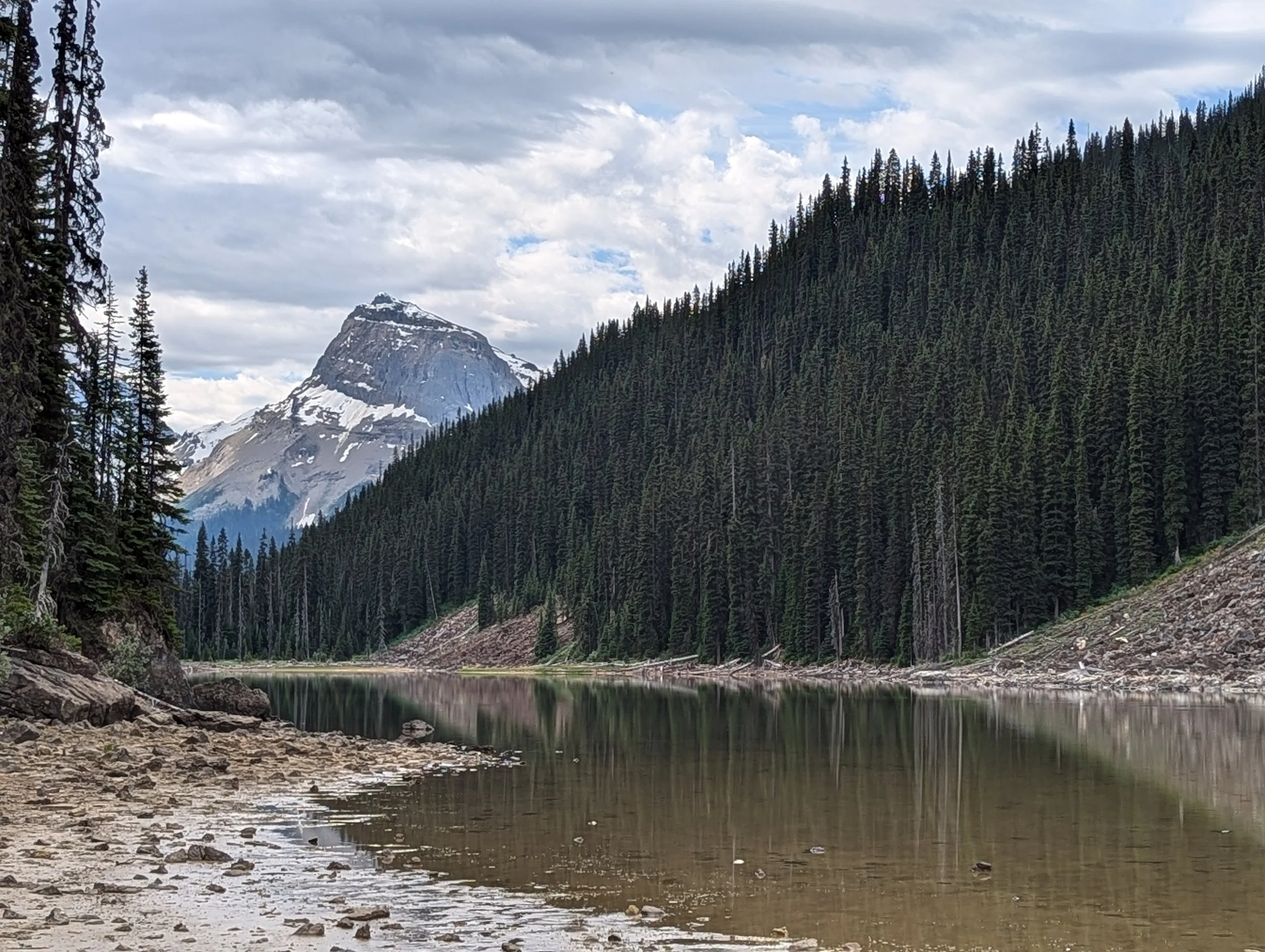 A mountain with snow at the top behind a dense forest of evergreen trees, a lake in the foreground, and a cloudy sky overhead.