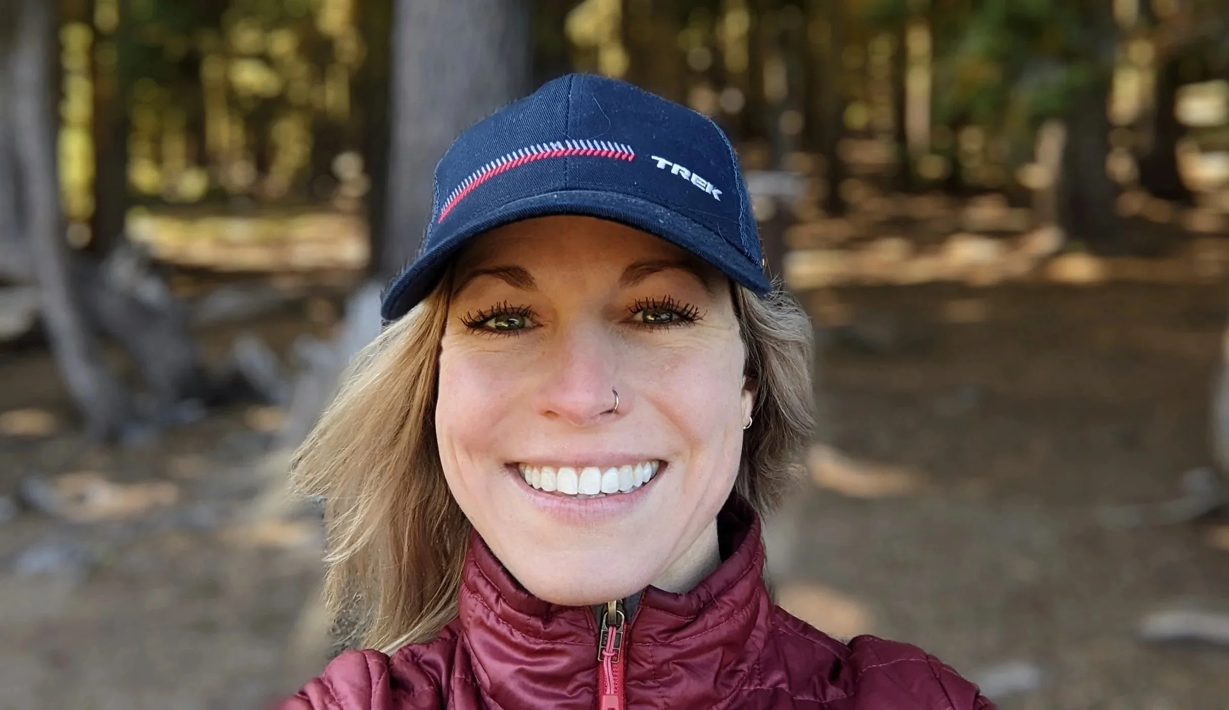 A woman smiling outdoors, wearing a blue cap with red and white accents and the word 'TREK' on it, and a red puffy jacket, with trees and a forest background.