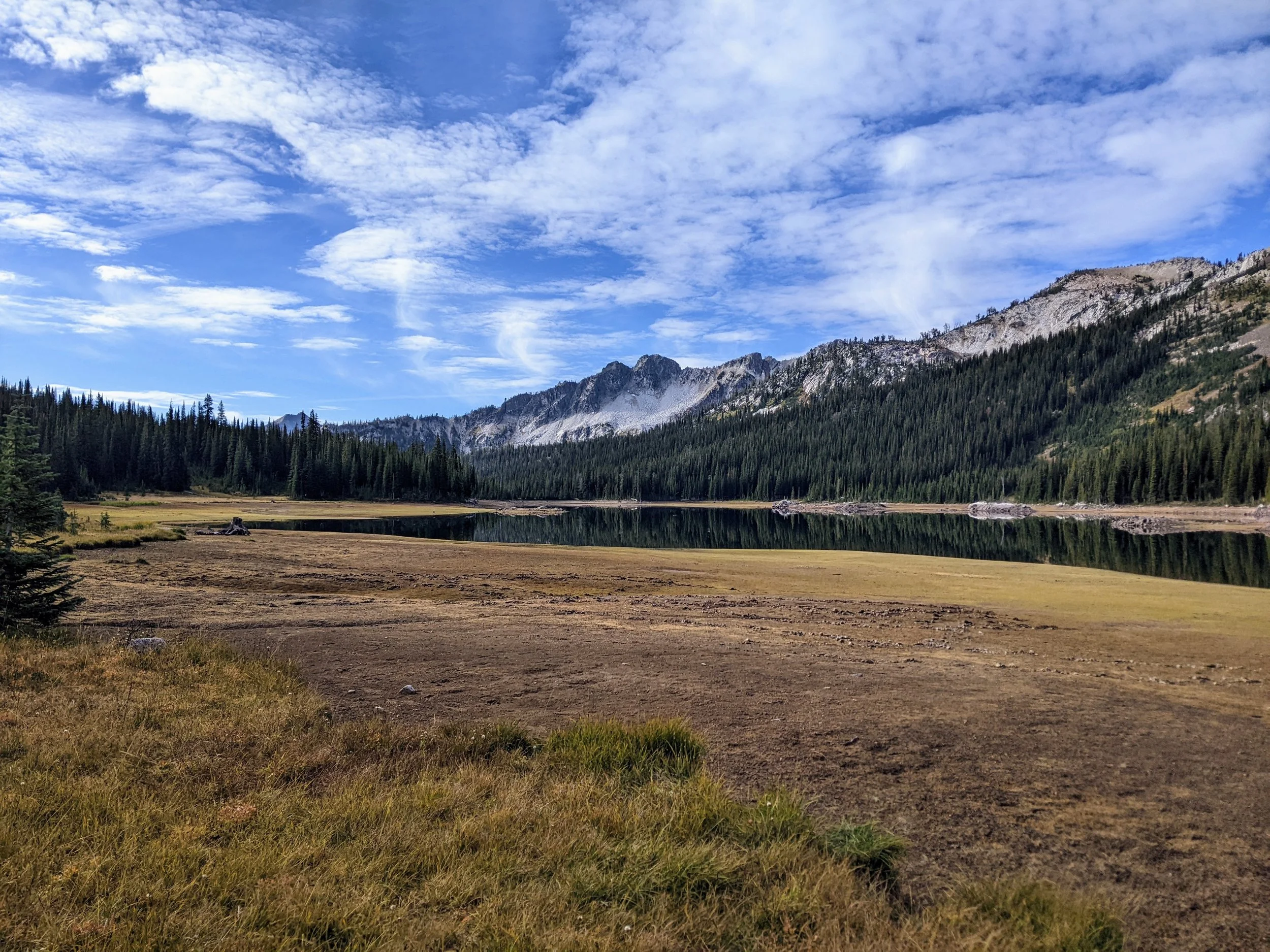 A landscape photo showing a pine forest at the foot of rocky mountains under a partly cloudy sky, with a body of water reflecting the scene.