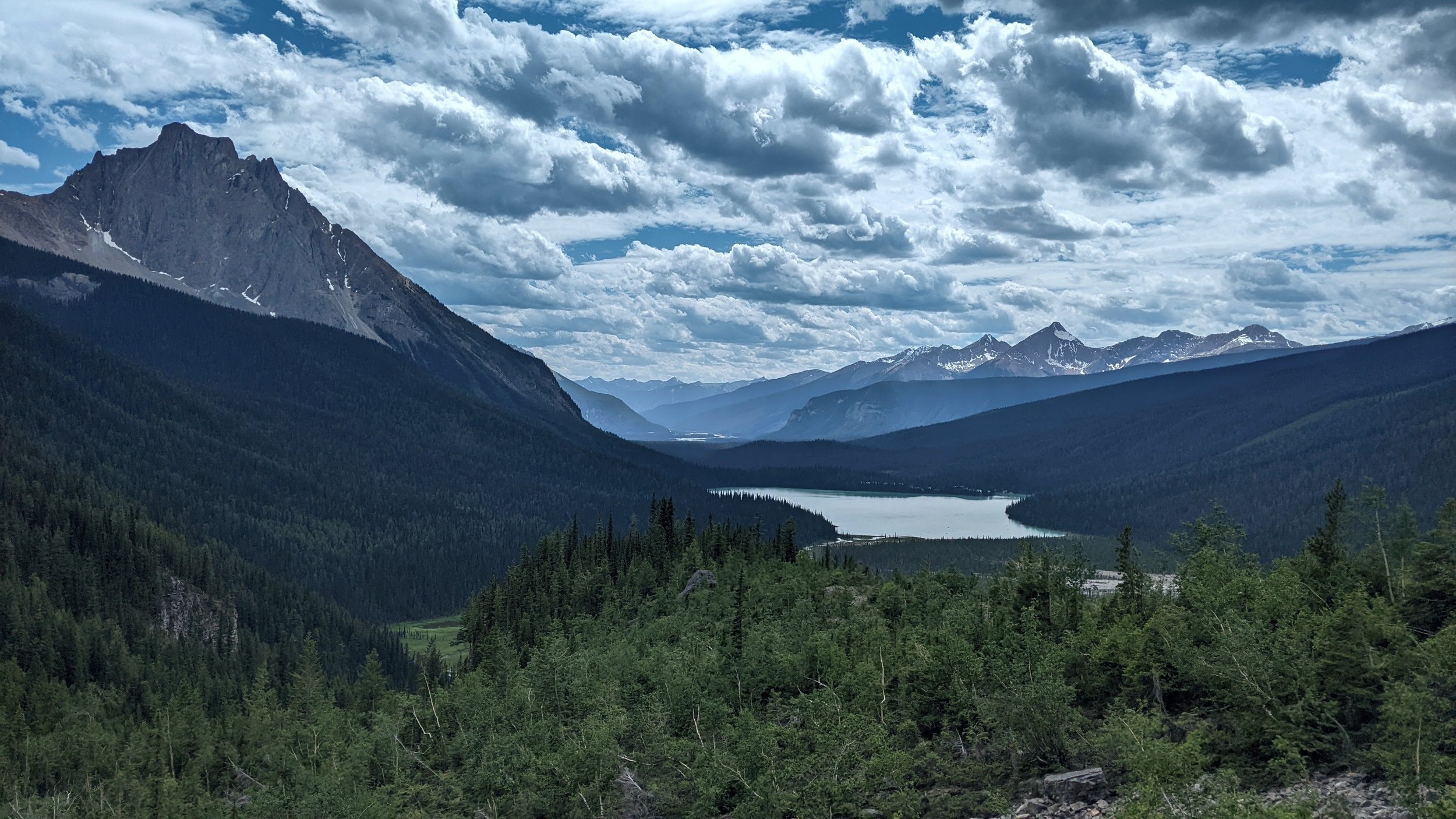 A scenic view of a mountainous landscape with snow-capped peaks, a lake in the valley, and dense green forests in the foreground under a partly cloudy sky.