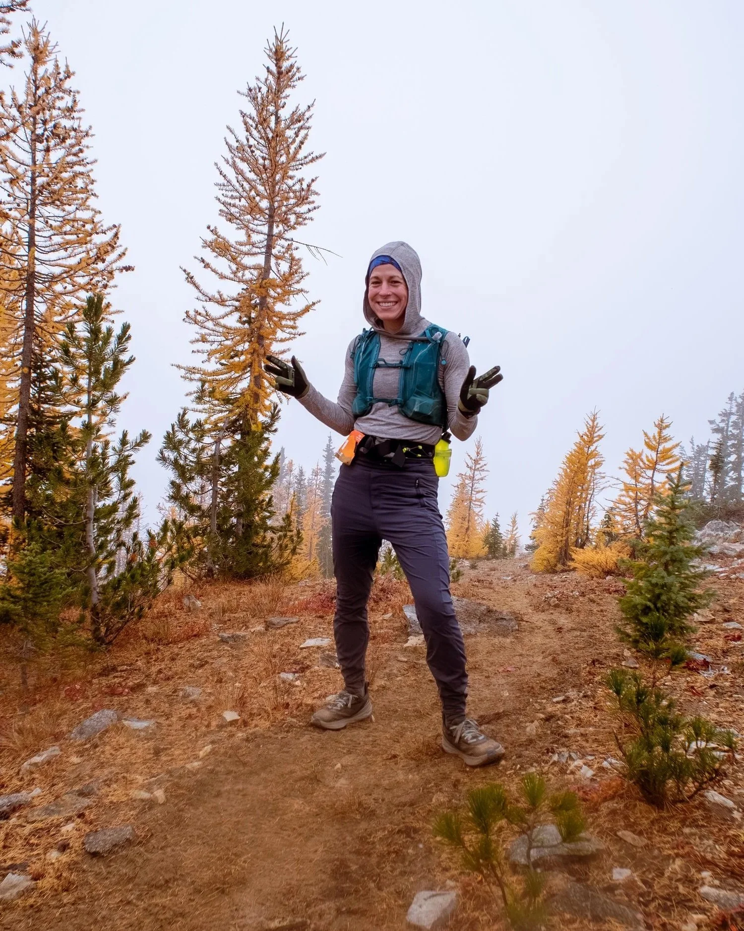 A woman hiking outdoors in a forested area with tall, yellow-orange trees, wearing outdoor gear and smiling at the camera.