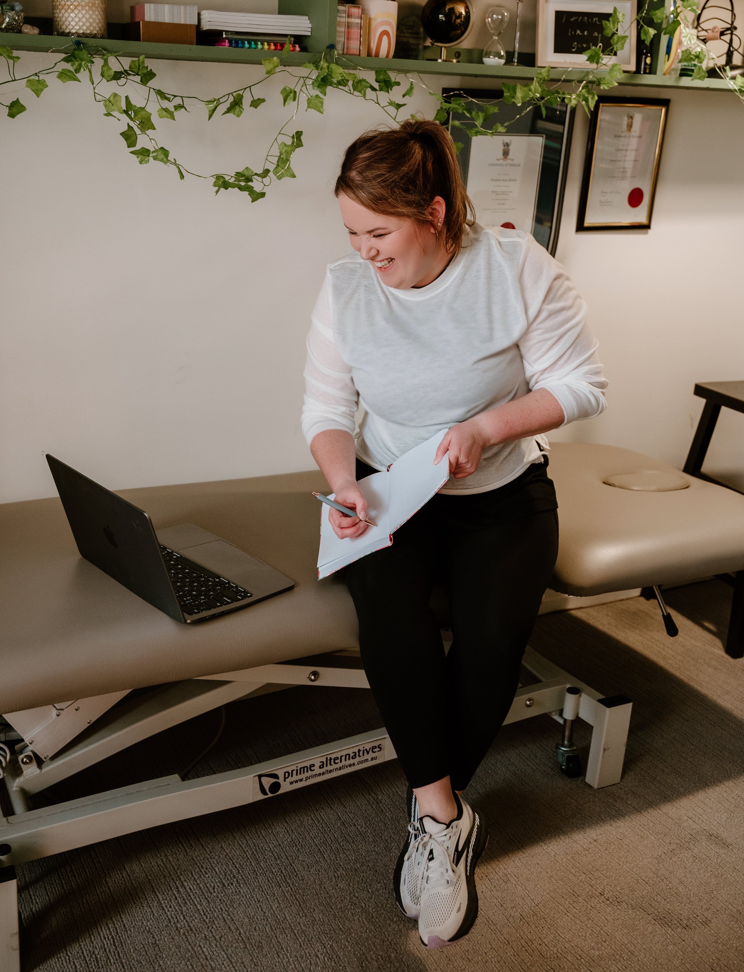 A woman in casual clothes, including a white long-sleeve shirt, black pants, and sneakers, sitting on a therapy or examination table in an office or clinic. She is smiling while holding a notebook and a pen, with a laptop in front of her. The background has shelves with books, certificates, and decorative items, and green ivy hanging overhead.