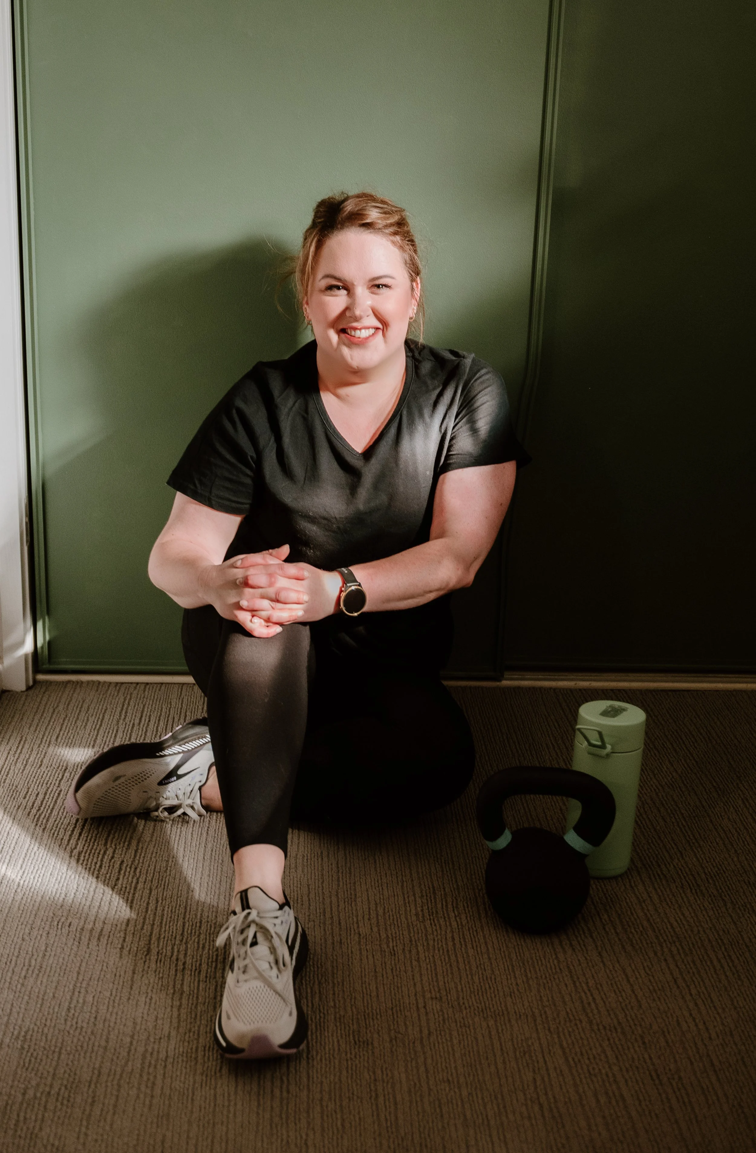A woman in workout clothes kneeling on the floor, smiling, with a kettlebell, water bottle, and workout towel nearby.